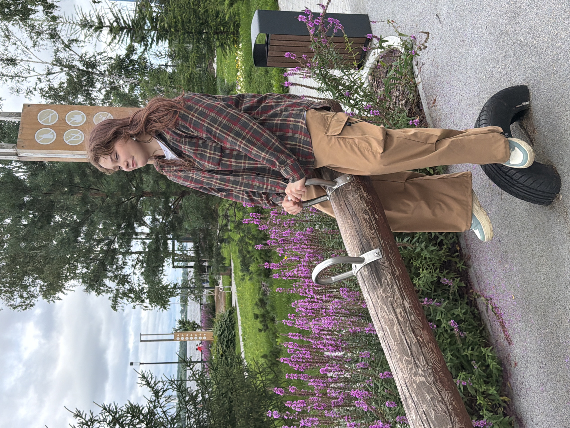 A young woman with shoulder-length reddish-brown hair is sitting on a seesaw in a park. She is wearing a red, green, and beige plaid flannel shirt over a white t-shirt, and loose-fitting tan cargo pants. Her hands are gripping the metal handles of the seesaw, and her feet are resting on a black tire that serves as the seesaw's base. She is looking to her left with a slight smile.
The seesaw is made of a large, rough-hewn log. Behind the woman, a row of tall, vibrant purple flowers lines the pathway. To the right of the seesaw, a dark gray trash receptacle with wooden slats is visible. Further back, a tall, light-colored structure with circular icons is positioned near a body of water, likely the Kama River, given the location in Perm, Russia. Evergreen trees and deciduous trees are scattered throughout the park, and the sky is overcast with gray clouds. The ground is paved with a textured gray surface.