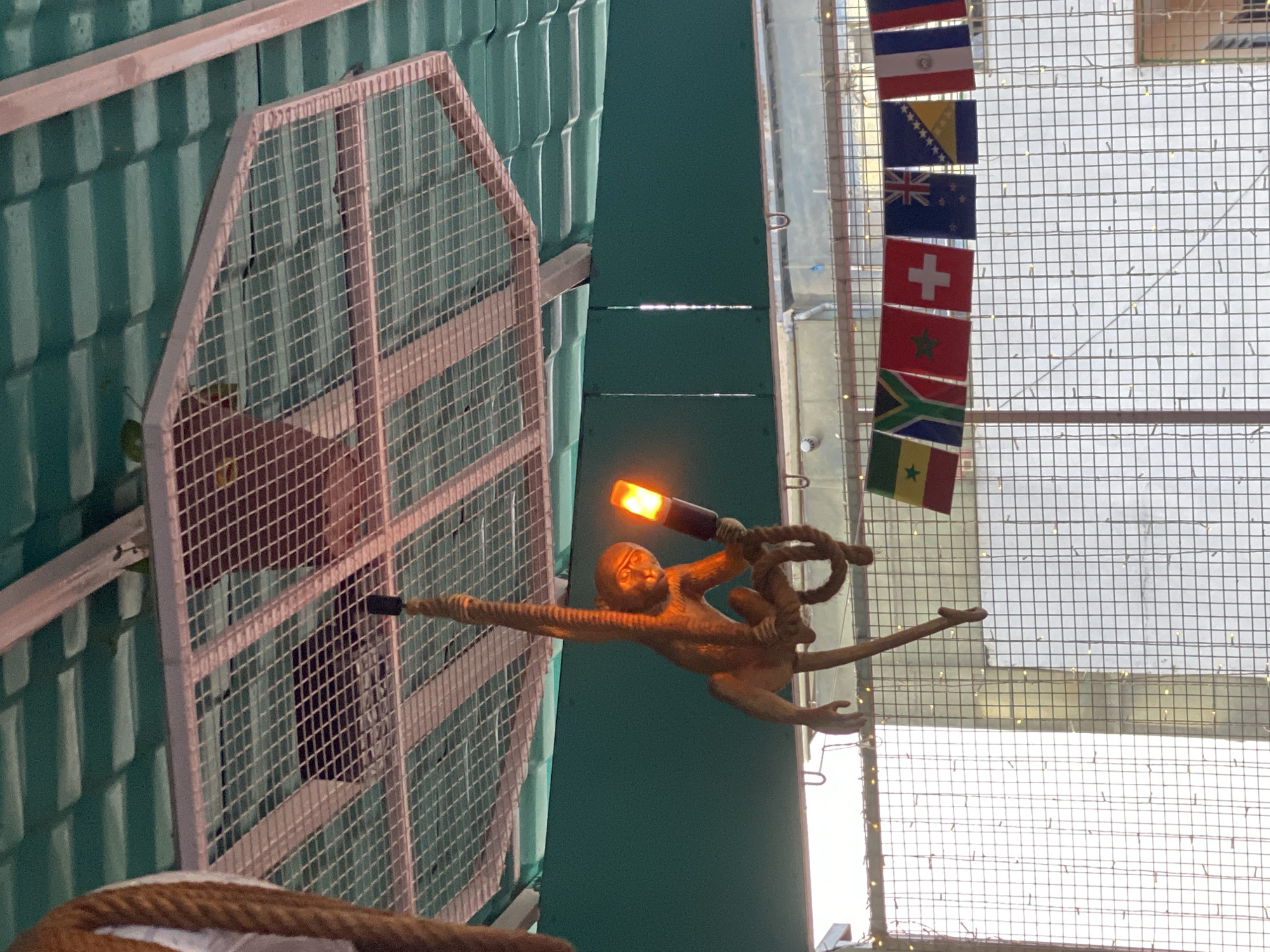 A decorative, tan-colored monkey figurine hangs by a rope from a metal cage structure suspended from a green, corrugated roof. The monkey is holding a bright orange, cylindrical object that emits light. Below the monkey, a string of small, yellow lights is visible, and a row of national flags are displayed on a wire. The flags appear to represent various countries, including Senegal, South Africa, and Switzerland. The background shows a wire mesh structure and bright, possibly natural, light. The scene appears to be outdoors, under a covered area, possibly a market or a tourist attraction.