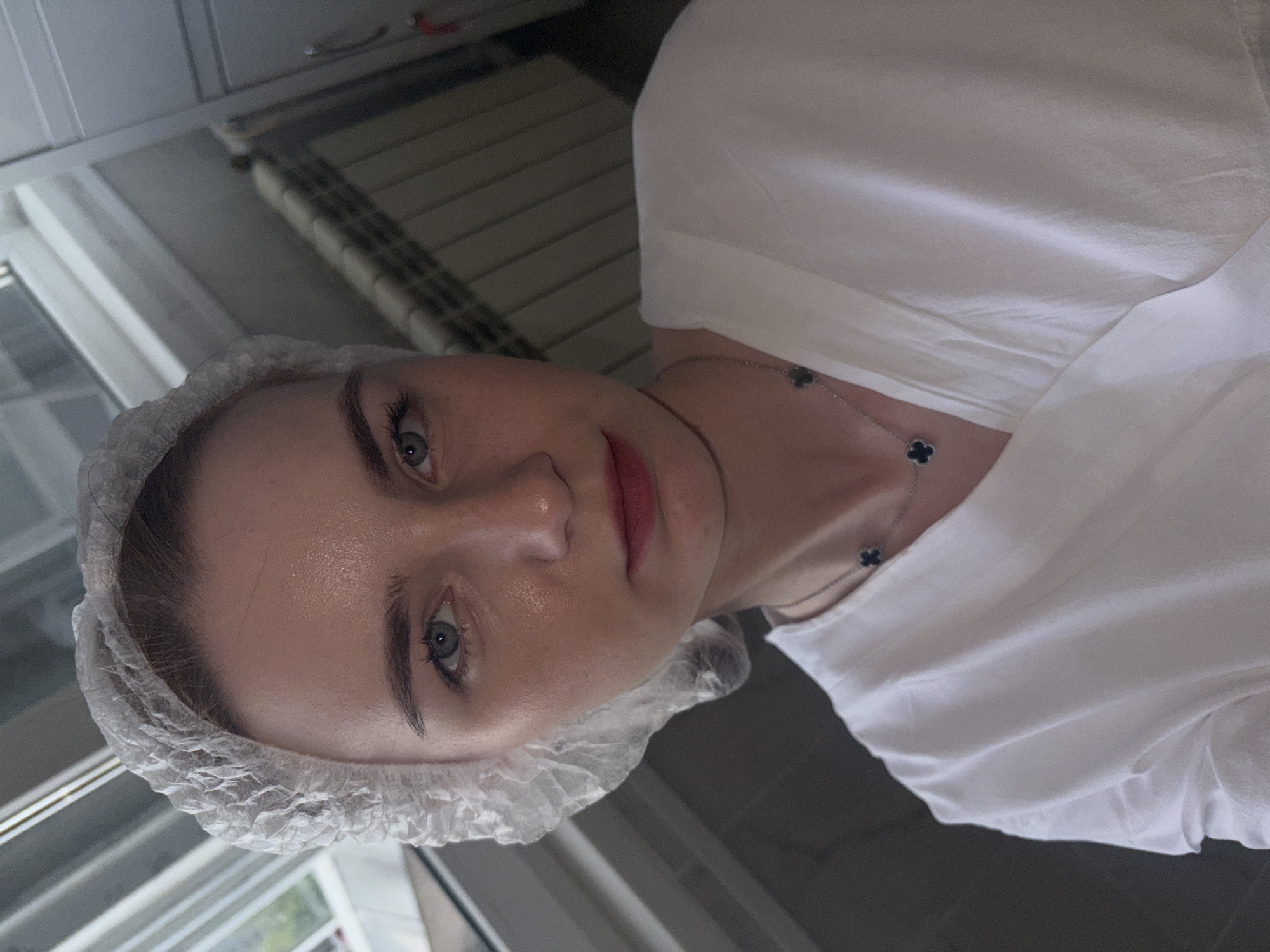A young woman, wearing a white disposable hair net and a white v-neck top with a silver necklace featuring dark clover-shaped pendants, looks directly at the camera. Her eyes are light blue, and her lips are painted with a reddish lipstick. Her skin appears dewy. The background is out of focus, showing what appears to be a window with horizontal blinds on the left, and a radiator with vertical slats on the right, suggesting an indoor setting that could be a home or a professional environment. The lighting is soft, casting a warm glow on her face.