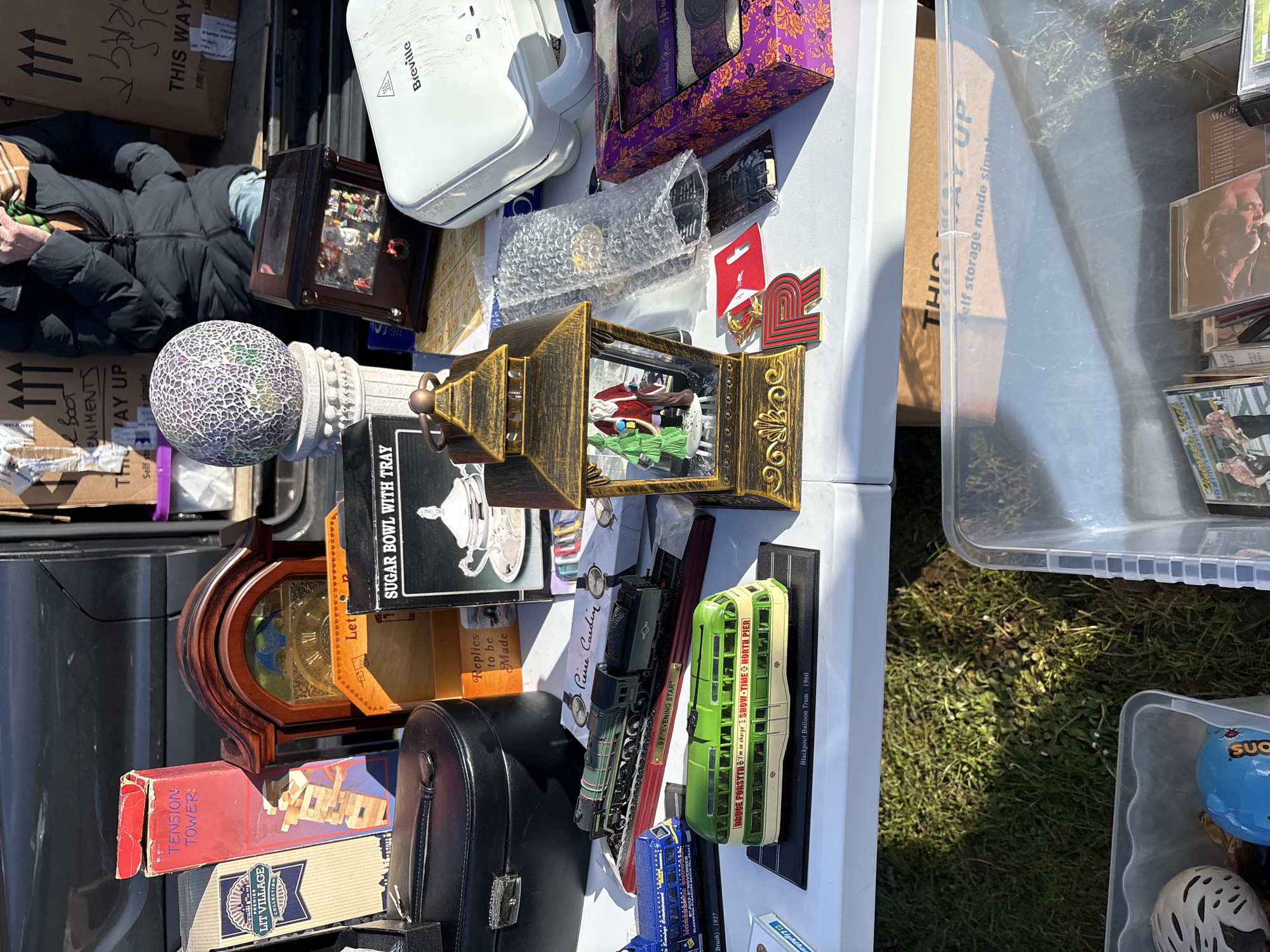 This image shows a collection of various items for sale, likely at a flea market or yard sale in Halton, United Kingdom. The items are displayed on a white table. The collection includes a Breville panini press, a purple decorative box, various smaller decorative boxes, an ornate gold-colored lantern with figurines inside, a "Sugar Bowl with Tray" box, a wooden clock, a "Tension Towers" game box, a "Lit Village" collection box, toy train models, a "Pierre Cardin" box, a "Bruce Forsyth - I'm in charge" Blackpool Balloon Tram model, CDs, and a clear plastic storage bin. A person is seen in the background near some cardboard boxes, likely the seller. The background also shows patches of grass.