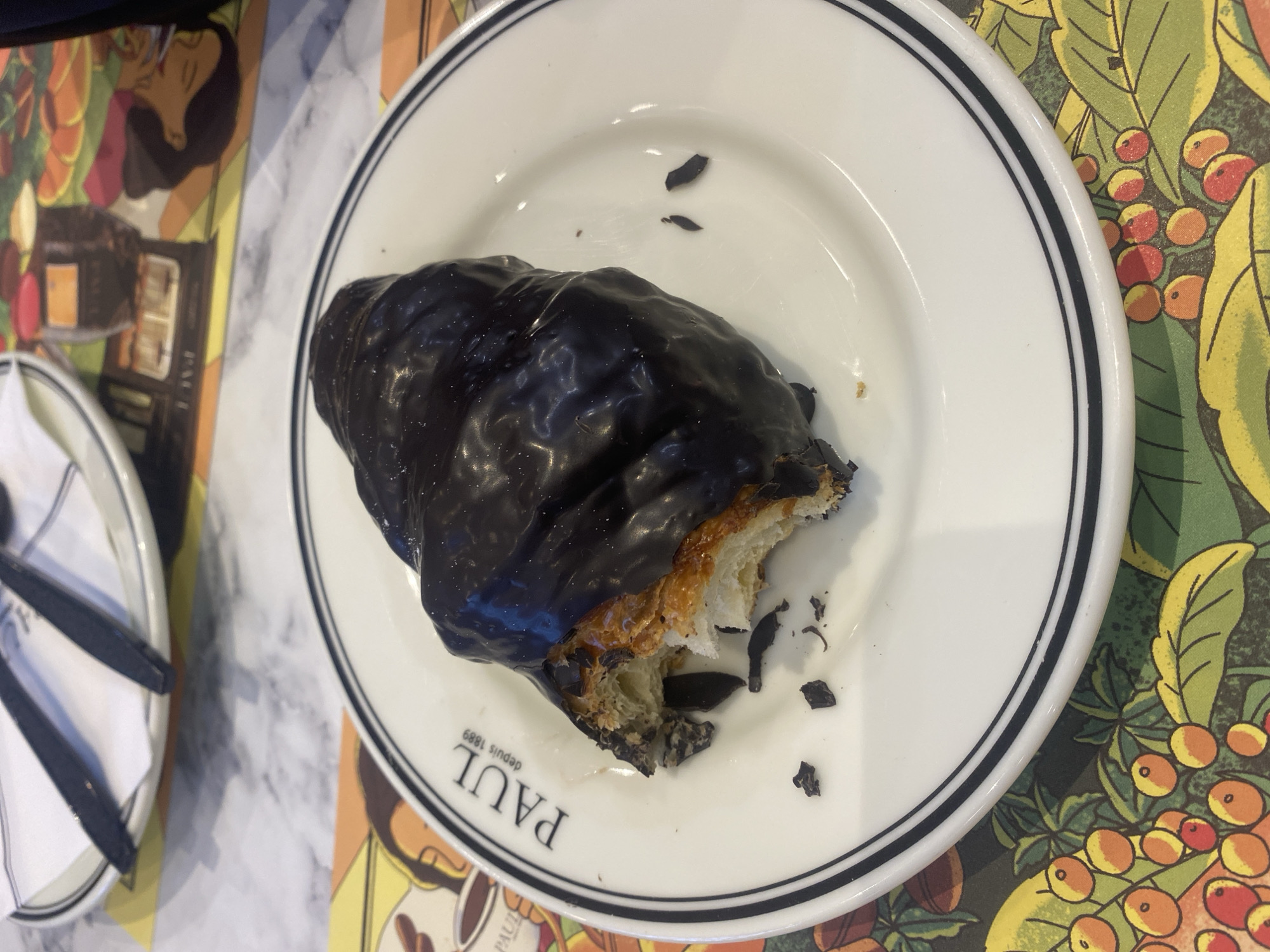 A chocolate-covered croissant on a white plate with a black rim sits on a table with a patterned placemat. The croissant is partially eaten, revealing the inner layers. The plate has the word "PAUL" and "depuis 1889" printed on it. A partially visible plate with silverware is to the left of the main plate. The location is Nong Prue Subdistrict, Thailand.