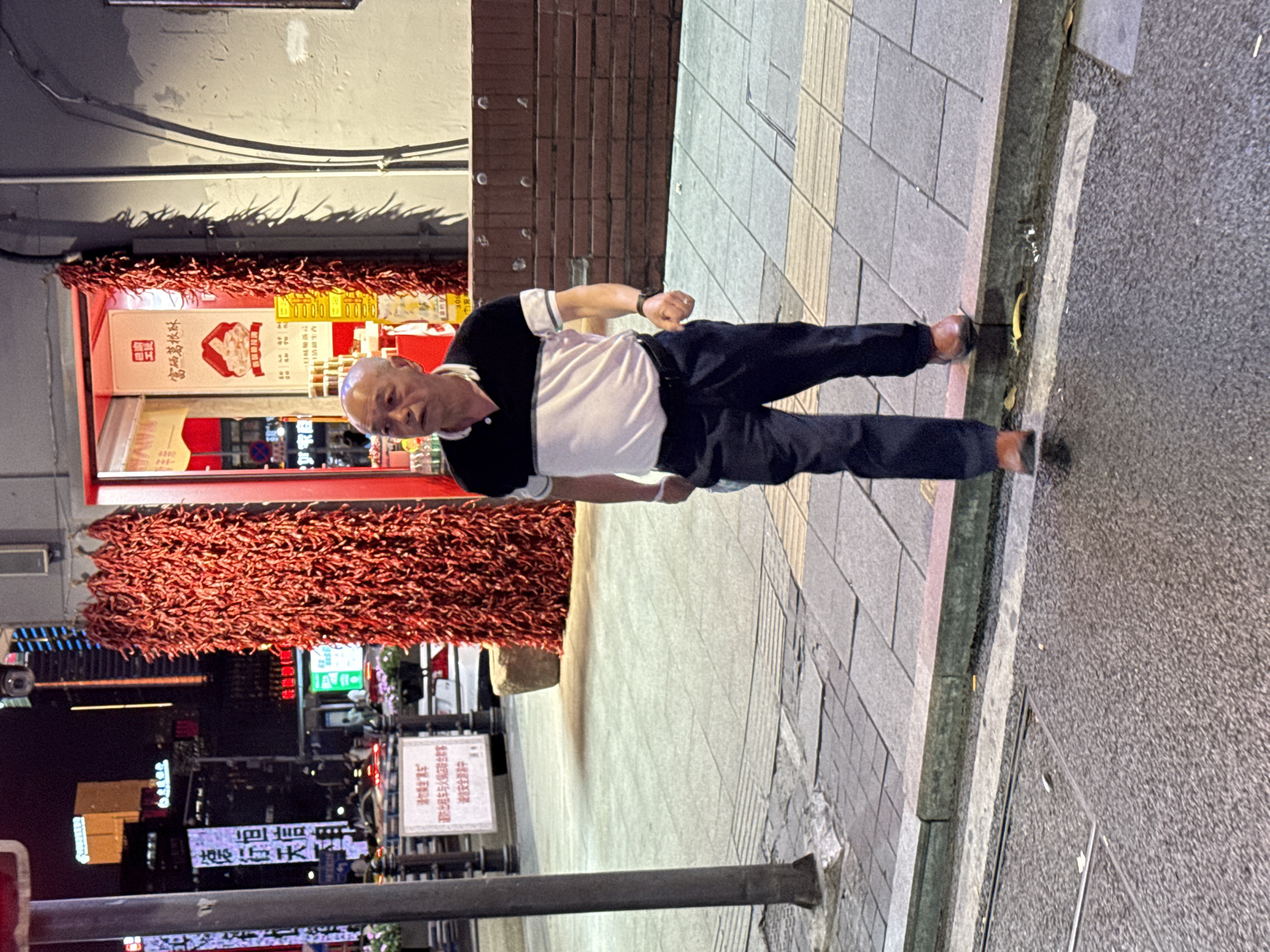 The image is taken at night in Yuzhong District, China. An older man stands on a sidewalk. He is wearing a black and white shirt and dark pants. Behind him is a storefront with red decorations and a sign in Chinese. Nighttime lights from buildings are visible in the background.