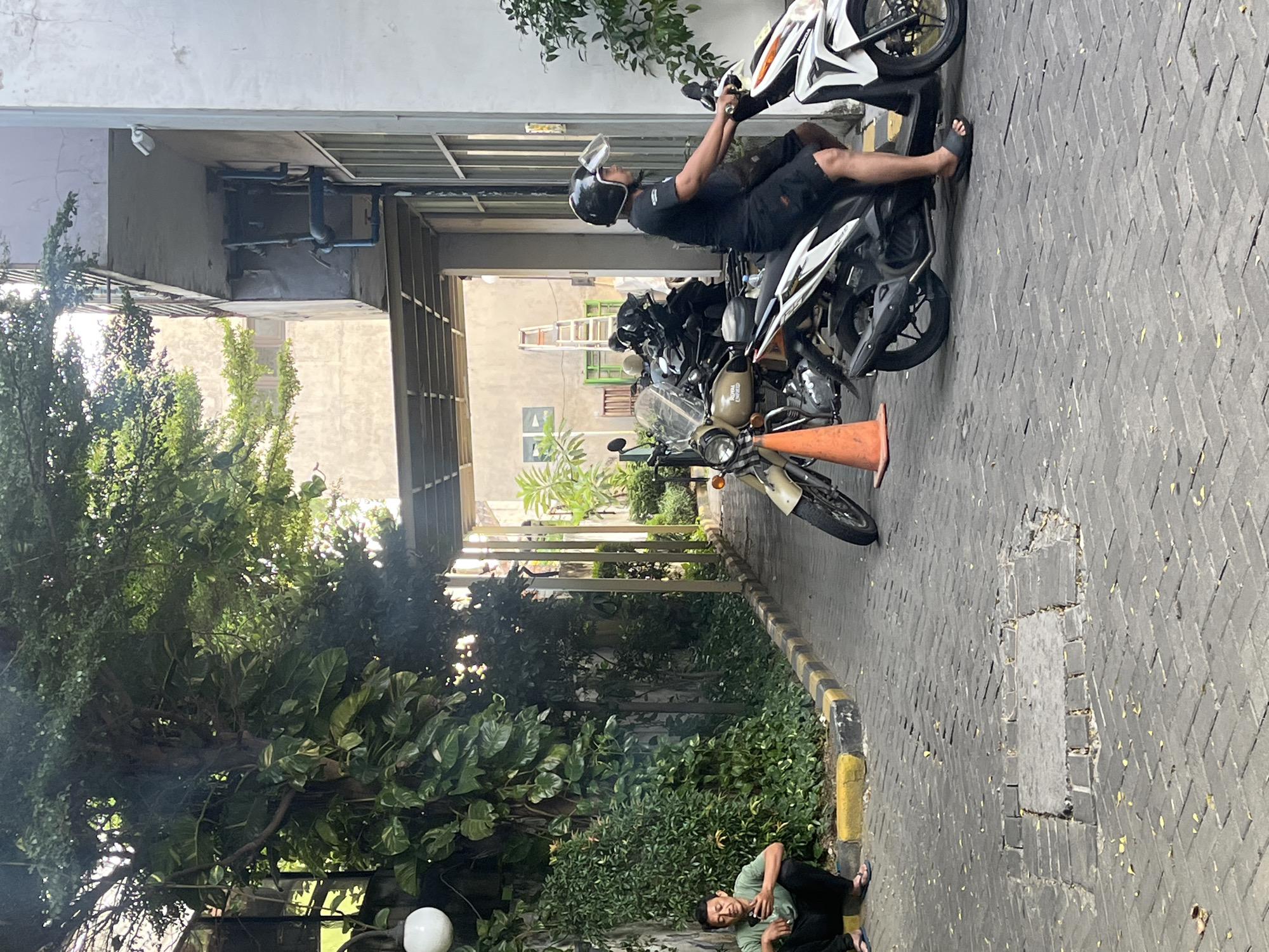 In Gapurosukolilo, Indonesia, a man in a green shirt and black pants is sitting on a yellow and black striped curb next to lush green bushes, looking at his phone. Adjacent to him are several motorcycles parked. In the foreground, a person wearing a black t-shirt, black shorts, and black sandals is riding a white and black motorcycle. Another beige motorcycle is parked to the left of the person riding the white and black motorcycle. The ground is paved with interlocking brick pavers, and fallen yellow leaves are scattered across the surface. The scene appears to be an outdoor area, possibly a street or a parking lot, with a building and trees visible in the background.