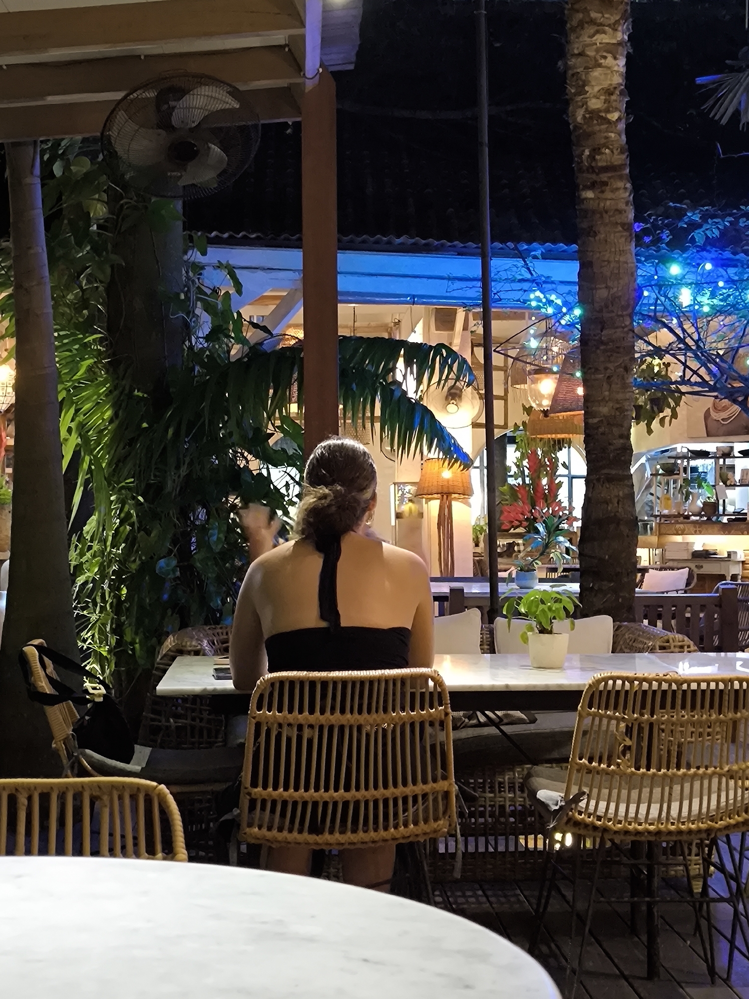 This image depicts an evening scene at an open-air or semi-outdoor restaurant in North Kuta, Indonesia. The primary subject is a woman, seen from behind, seated at a white marble-top table. She has dark hair tied back in a bun and is wearing a black strapless top. She occupies a natural wicker chair, with another identical chair positioned to her left.
The setting is characterized by a tropical ambiance, featuring prominent palm trees and lush green foliage that line the dining area. The scene is illuminated by artificial lighting, indicating nighttime. Overhead, a black ceiling fan provides air circulation. The background features strung blue fairy lights, adding a festive glow, while warm pendant lights and a tall, woven floor lamp contribute to the cozy atmosphere.
On the table in front of the woman, a small potted green plant is visible. The venue is furnished with numerous other white marble tables and wicker chairs, some with white cushions. A notable detail, revealed on the back of a wicker chair to the right, is a small lizard. The overall impression is one of a relaxed and inviting tropical dining or social setting. No text is visible in the image.