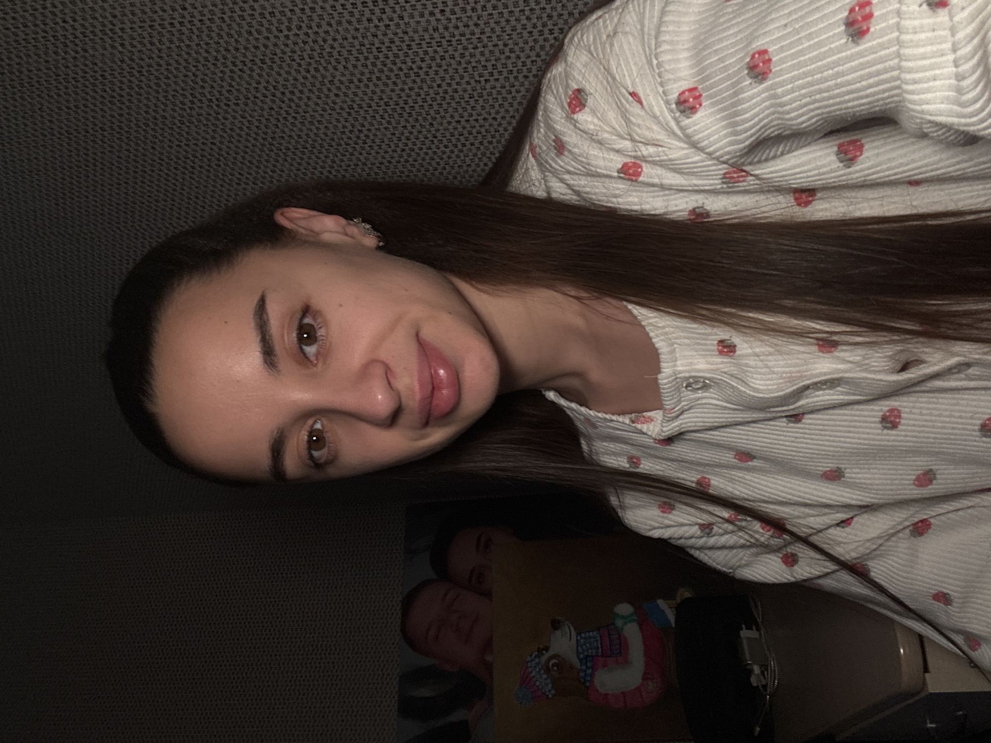 A woman with long, dark hair is indoors. She is wearing a light-colored, button-down shirt with red strawberry patterns. The background is dark and textured. Below her, there's an image of two people, with one appearing to be a child holding a stuffed animal. The location is Fadeevskii, Russia.
