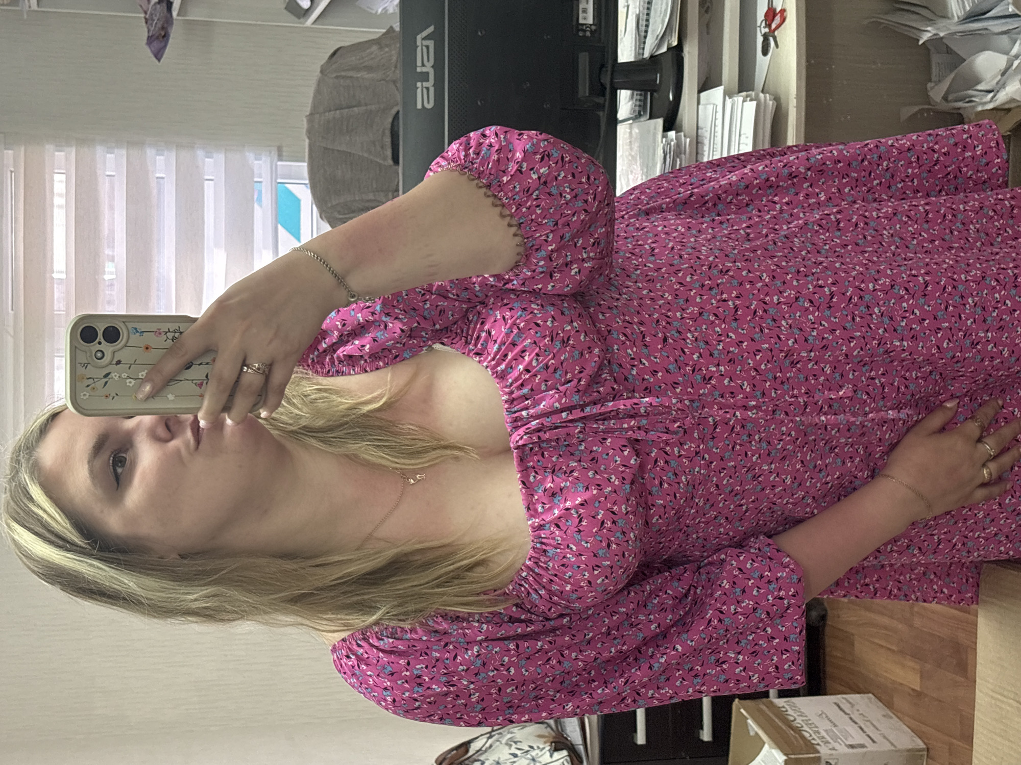 A woman with blonde hair is taking a selfie. She is wearing a pink dress with a floral pattern. She is holding a phone in her left hand and is standing in front of a window with blinds. There is a computer monitor and some papers in the background. The location is unclear. There is no mention of Noviy, Russia.