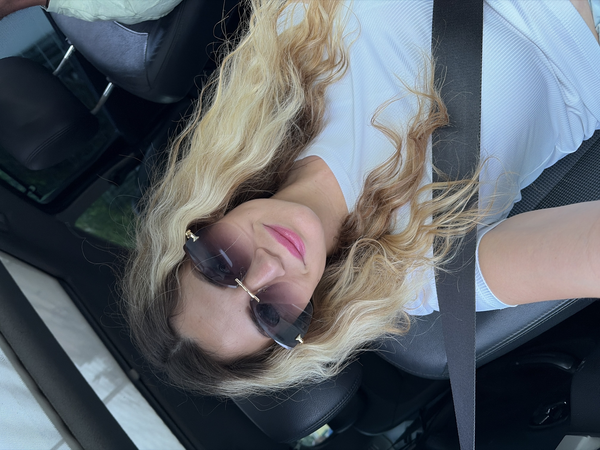 In Budva, Montenegro, a woman with long, wavy blonde hair is pictured inside a car, wearing sunglasses and a white ribbed t-shirt. She is seated and appears to be wearing a seatbelt across her chest. The interior of the car is visible, with dark leather seats and a sunroof above. The lighting suggests it is daytime, and the view through the windows indicates an outdoor setting.