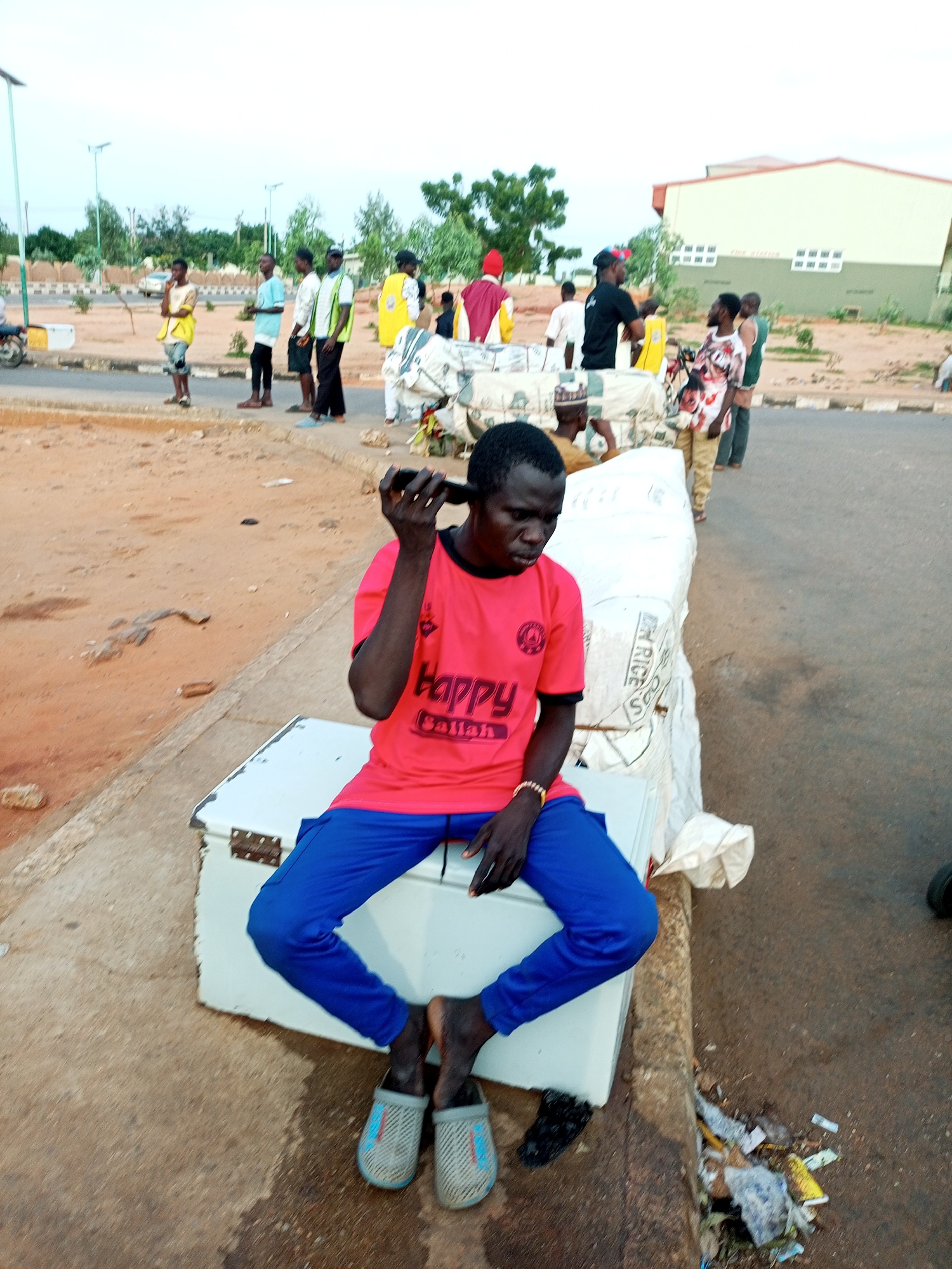 In an outdoor setting in the city of Unknown, Unknown, a young man is seated on a white rectangular object, possibly a cooler or storage box. He is wearing a vibrant red t-shirt with black lettering and blue pants. His gaze is directed downwards, and he holds a dark object, likely a mobile phone, to his ear. His left hand rests on his lap, and he has a yellow beaded bracelet on his wrist. He is wearing light gray slip-on shoes with a blue and red pattern.
The ground around him is a mix of dirt and asphalt. To his left, a portion of the white object he is sitting on is visible, with what appears to be a light-colored sack or covering draped over it. In the background, several other people are standing, some wearing casual clothing and others in more formal or traditional attire, suggesting a public gathering or transit area. A building with a light yellow facade and a green base is visible in the distance. The sky is overcast. The scene appears to be a street or a roadside.