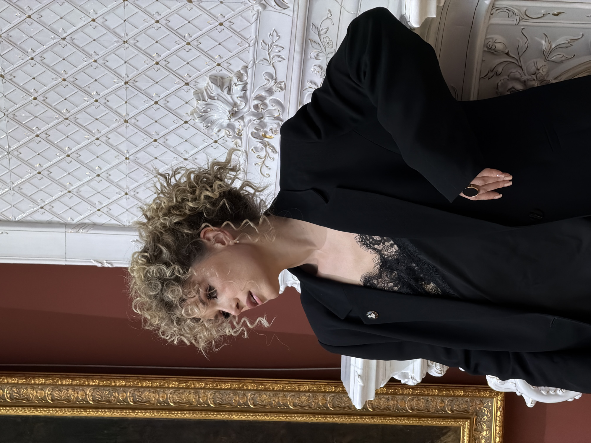 A woman with curly blonde hair is wearing a black blazer over a black lace top. She is indoors, possibly in a historical building or museum, as evidenced by the ornate, patterned ceiling, white decorative moldings, and a large framed painting with a gold frame. The wall behind her is a deep red. The woman is wearing a ring on her finger. The location is Kazan, Russia.