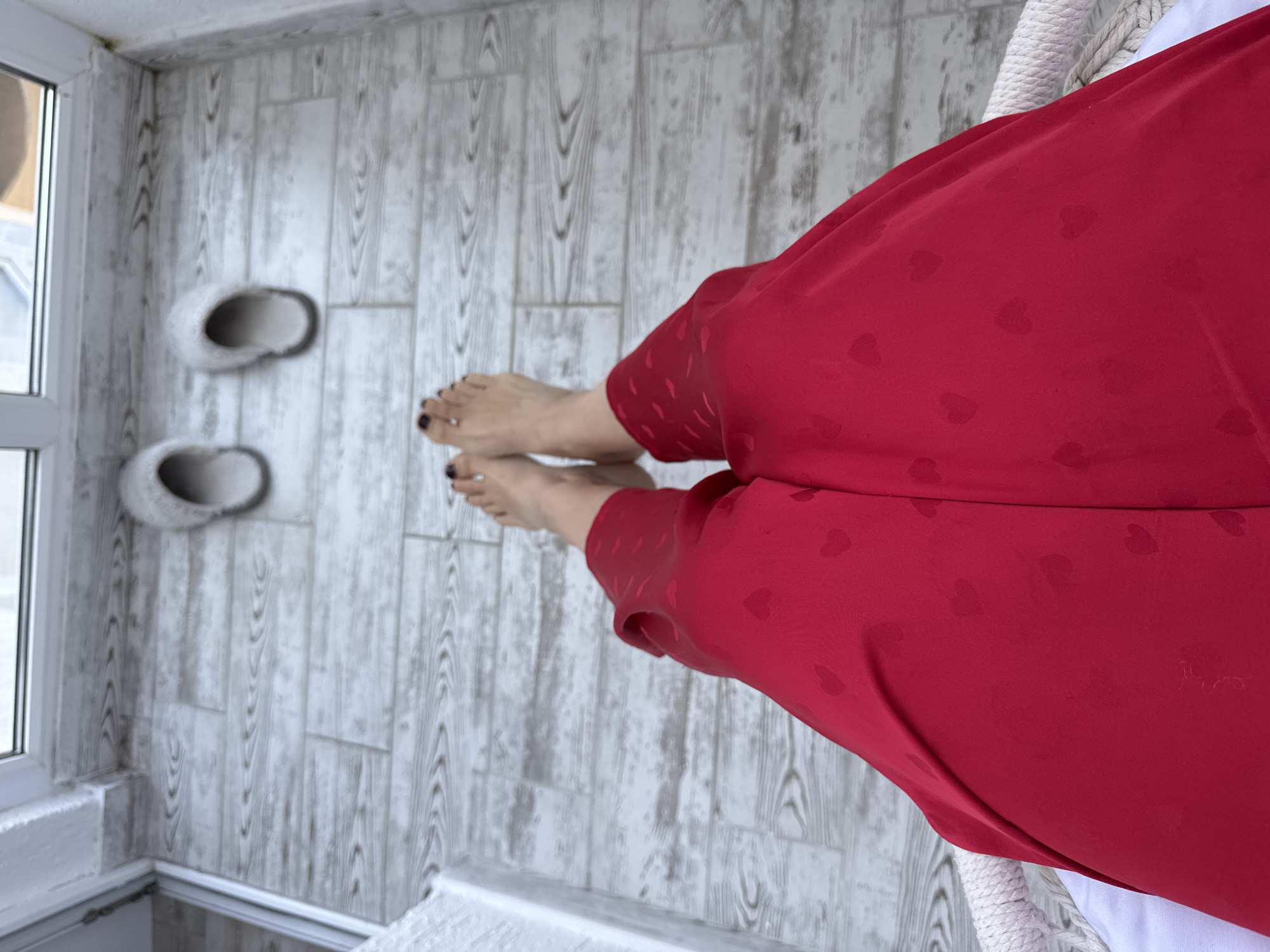 The image shows a pair of bare feet wearing dark nail polish, resting on a wooden-planked floor. The feet are adorned with red pants that have a subtle heart pattern. In the background, a pair of fluffy white slippers are placed on the floor. The scene suggests a relaxed indoor setting, potentially a balcony or patio. There is no indication of the specific location within the city of Sochi, Russia.
