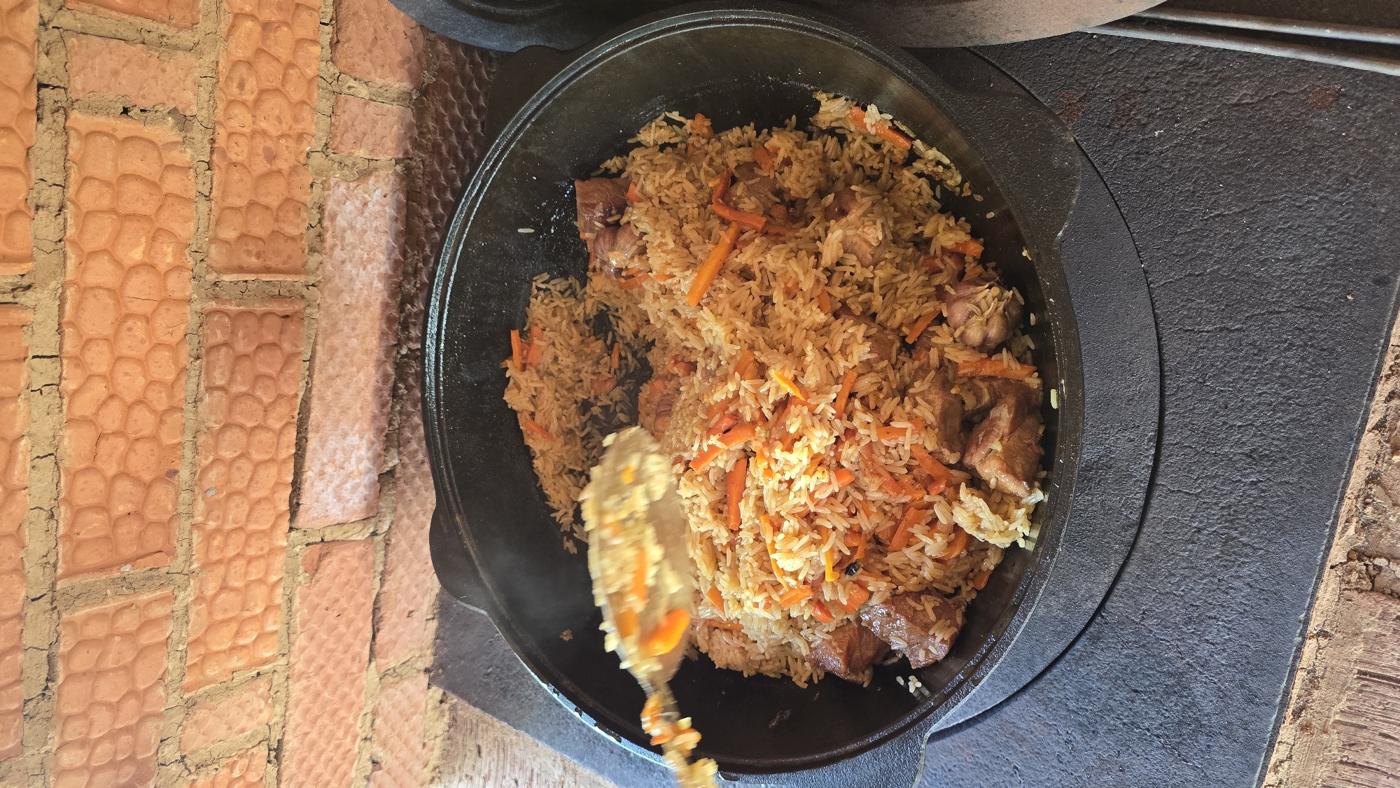 The image depicts a close-up view of a large, dark, cast-iron pot (Kazan) filled with a prepared rice dish, likely Plov, in Забегалово, Russia. The dish comprises light brown, long-grain rice, numerous bright orange julienned carrots, and several chunks of cooked meat. Several whole garlic cloves are also visible within the mixture. A metal slotted spoon is positioned on the left, actively lifting a portion of the rice and carrots from the pot, indicating stirring or serving. The pot rests on a heavy, dark grey, textured metallic surface, possibly a stovetop or a specialized outdoor cooking stove with a circular opening for the pot. In the background, a brick wall is visible, constructed from reddish-orange bricks featuring a distinctive, uneven, dimpled texture. The mortar between the bricks is a lighter, sandy brown color with visible cracks and inconsistencies. No people are discernible in the frame. The lighting suggests an outdoor or brightly lit indoor setting.