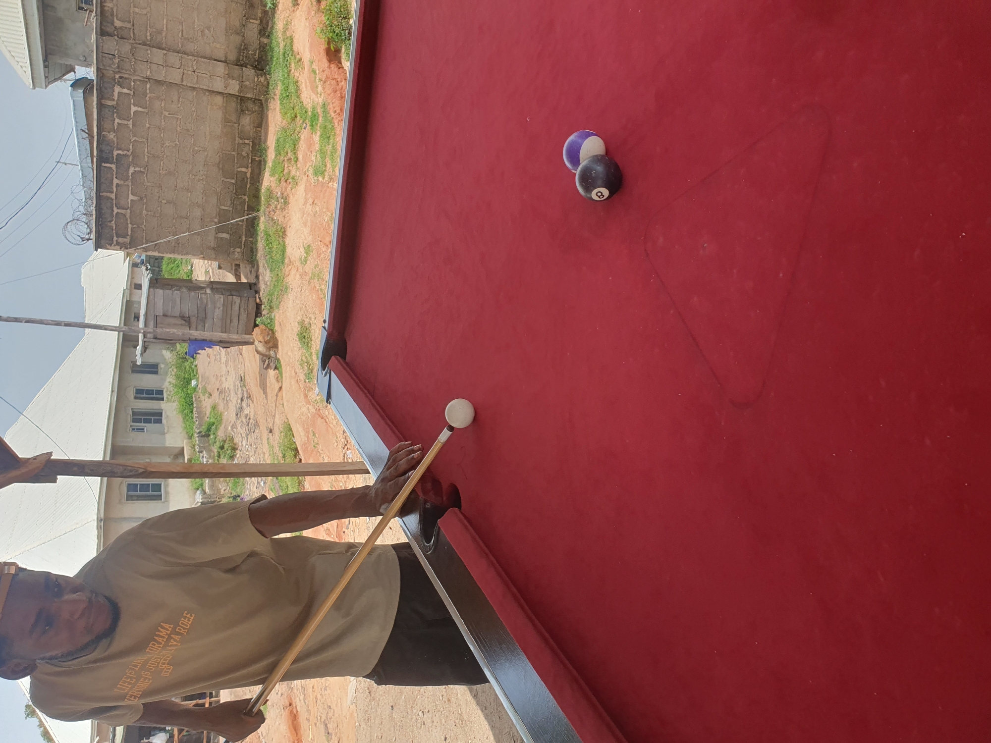 In Lokoja, Nigeria, a young Black man wearing a tan t-shirt with text reads "LIFE IS LIKE DRAMA EVERYONE IS JUST PLAY A ROLE" and a yellow cap is playing pool. He is holding a pool cue in his right hand, aimed at a white cue ball. The pool table has a red felt surface. Two balls are resting on the table: a black 8-ball and a purple ball. In the background, a dirt road is visible, along with a brick wall topped with barbed wire and some buildings. The sky is overcast.