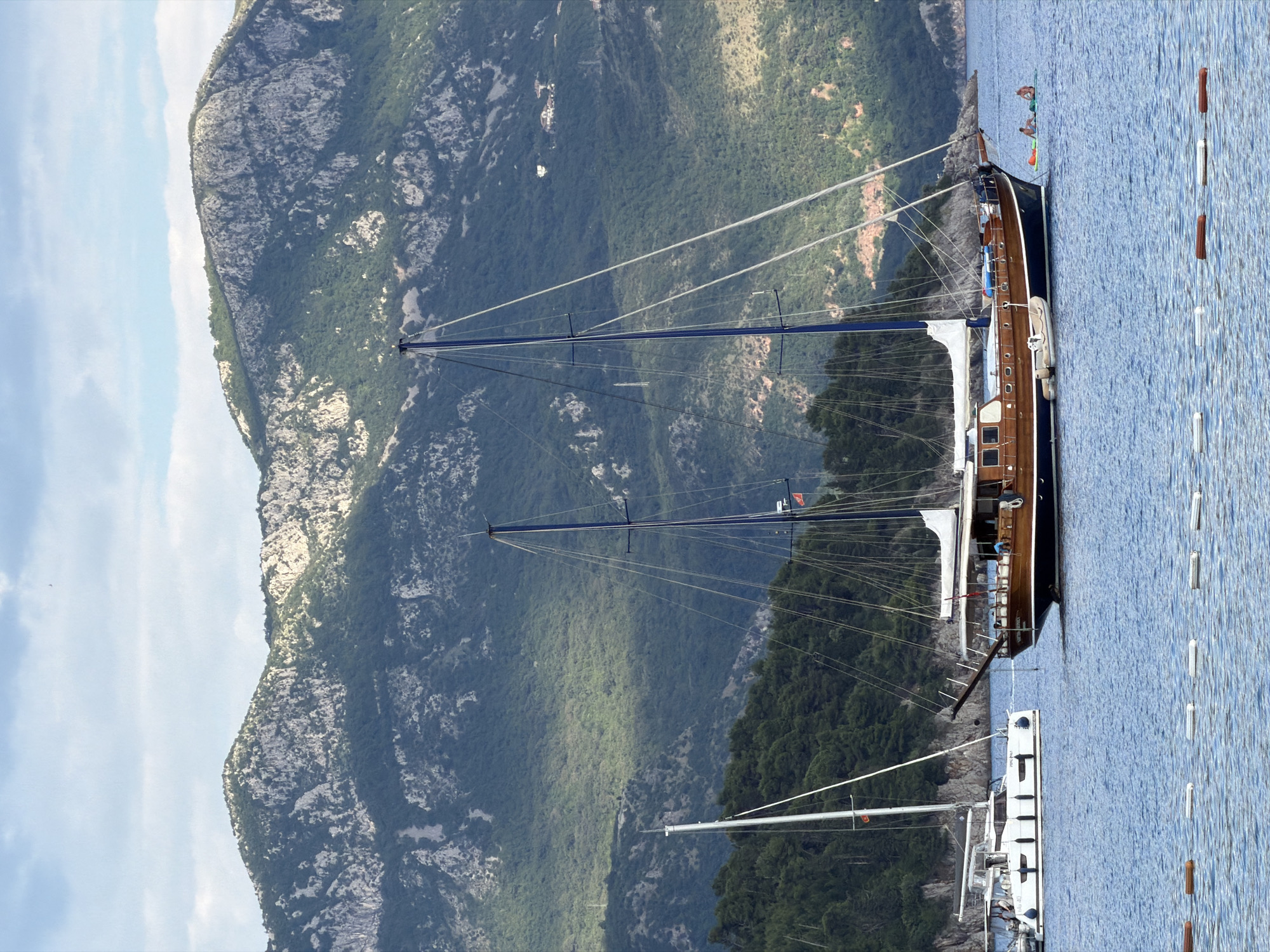 The scene is a view from the water in Budva, Montenegro. A large, two-masted sailing yacht is anchored close to shore. The yacht has a dark hull, wood trim, and sails. Near the yacht are two people on a stand-up paddleboard and a string of floating markers. A mountainous coastline covered in dense green vegetation and exposed rock forms the backdrop. The sky is partly cloudy.