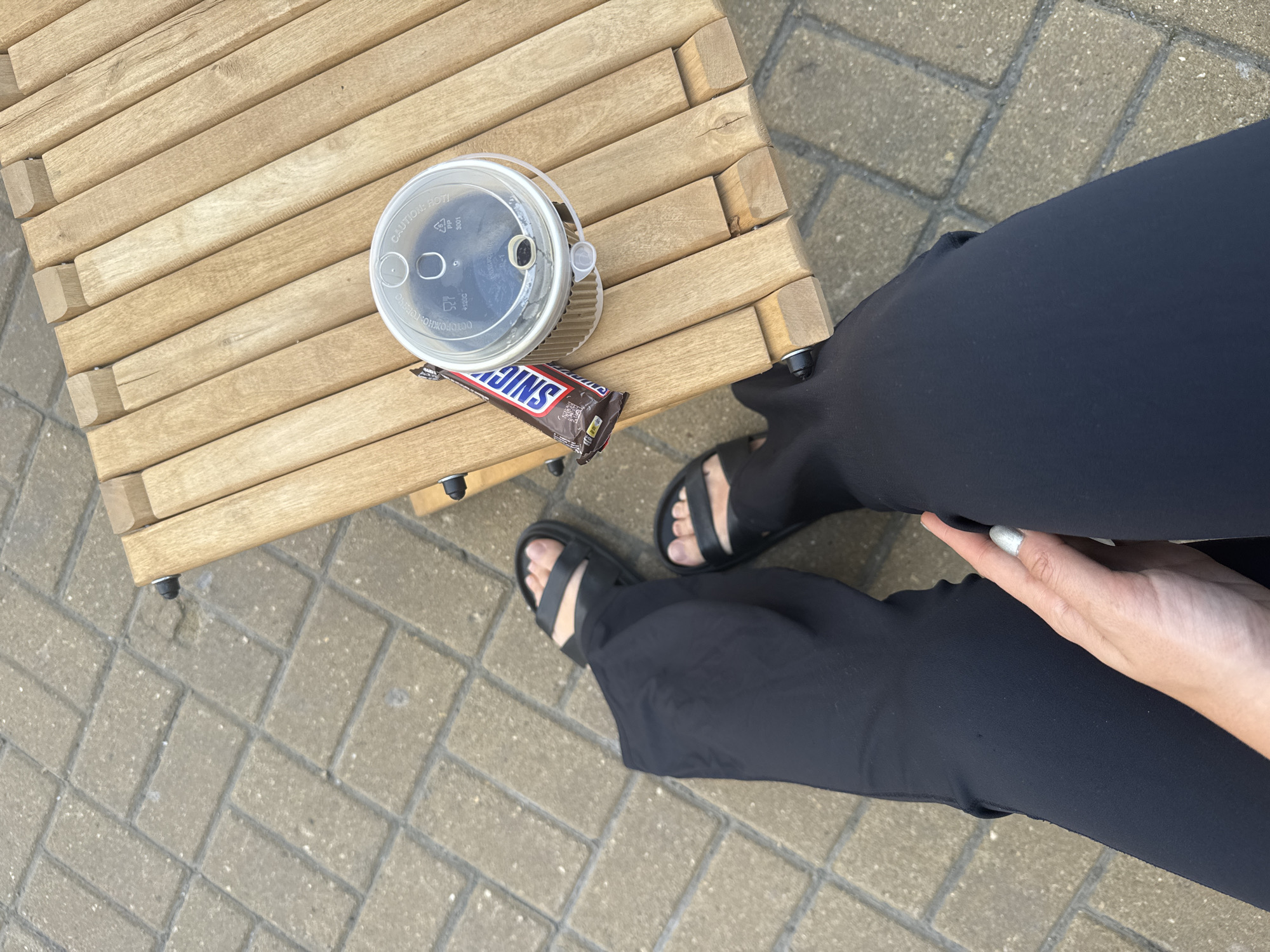 A person wearing black pants and black sandals sits on a wooden bench outdoors, with their feet resting on a paved ground. On the bench next to them are a cup of coffee and a Snickers bar. The scene appears to be a casual outdoor setting, possibly a cafe or park. There is no information in the image to suggest the location is Moscow, Russia.