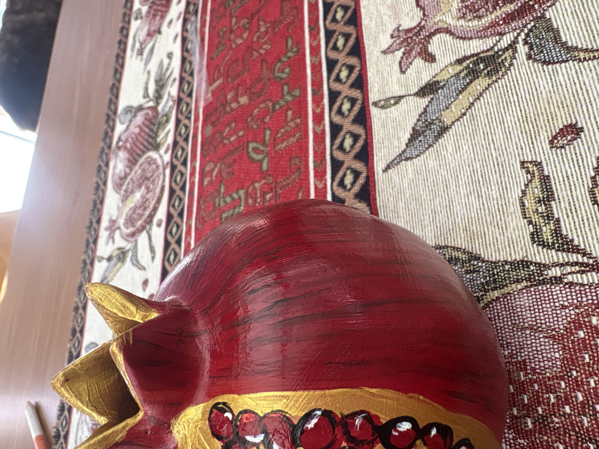 A close-up, angled shot shows a red, decorative pomegranate on a patterned table runner. The pomegranate is painted with gold accents and appears to be made of ceramic or a similar material. The table runner has intricate patterns in red, gold, black, and beige, featuring pomegranate motifs and geometric designs. A wooden surface, likely a table, is visible in the background, along with a blurred object that could be a pen and a dark, out-of-focus shape possibly representing a pet. The scene is indoors, with natural light suggesting a daytime setting. The location is not specified in the image or provided context.