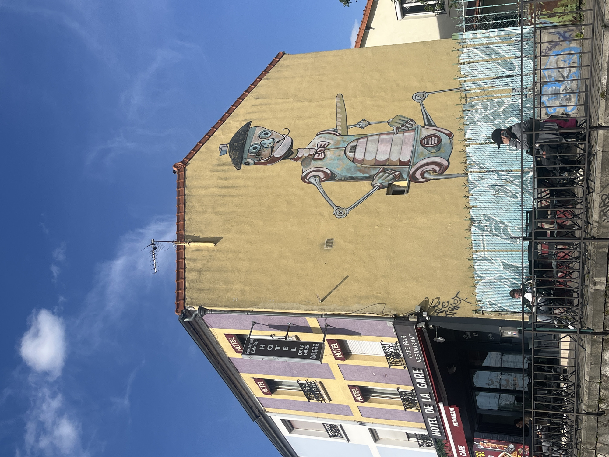 This media file captures a vibrant street scene in Vitry-sur-Seine, France, under a clear, bright blue sky with a few wispy clouds, indicating daytime.
The primary subject is a large, whimsical mural painted on the yellow side of a building. The artwork depicts a stylized, robot-like character with a human-like face, large eyes, and a hat. Its body and limbs appear mechanical, constructed from various metal components, rendered in shades of blue, grey, and brown.
Below this yellow building, another structure features a facade with alternating purple and yellow sections, dotted with windows adorned with black wrought-iron railings. This building houses commercial establishments, prominently featuring the "HOTEL DE LA GARE," which also advertises itself as a "Restaurant Café Bar." Several signs bear these names, with one displaying a phone number: "01 46 82 65 99."
In the foreground, behind black metal railings, people are visible sitting at outdoor tables, suggesting patrons of the "Café Bar Restaurant." One individual, wearing a black cap, is clearly seen seated at a table. Some graffiti is present on the lower part of the yellow wall, behind the outdoor seating area. An antenna is visible on the rooftop of the building with the mural.