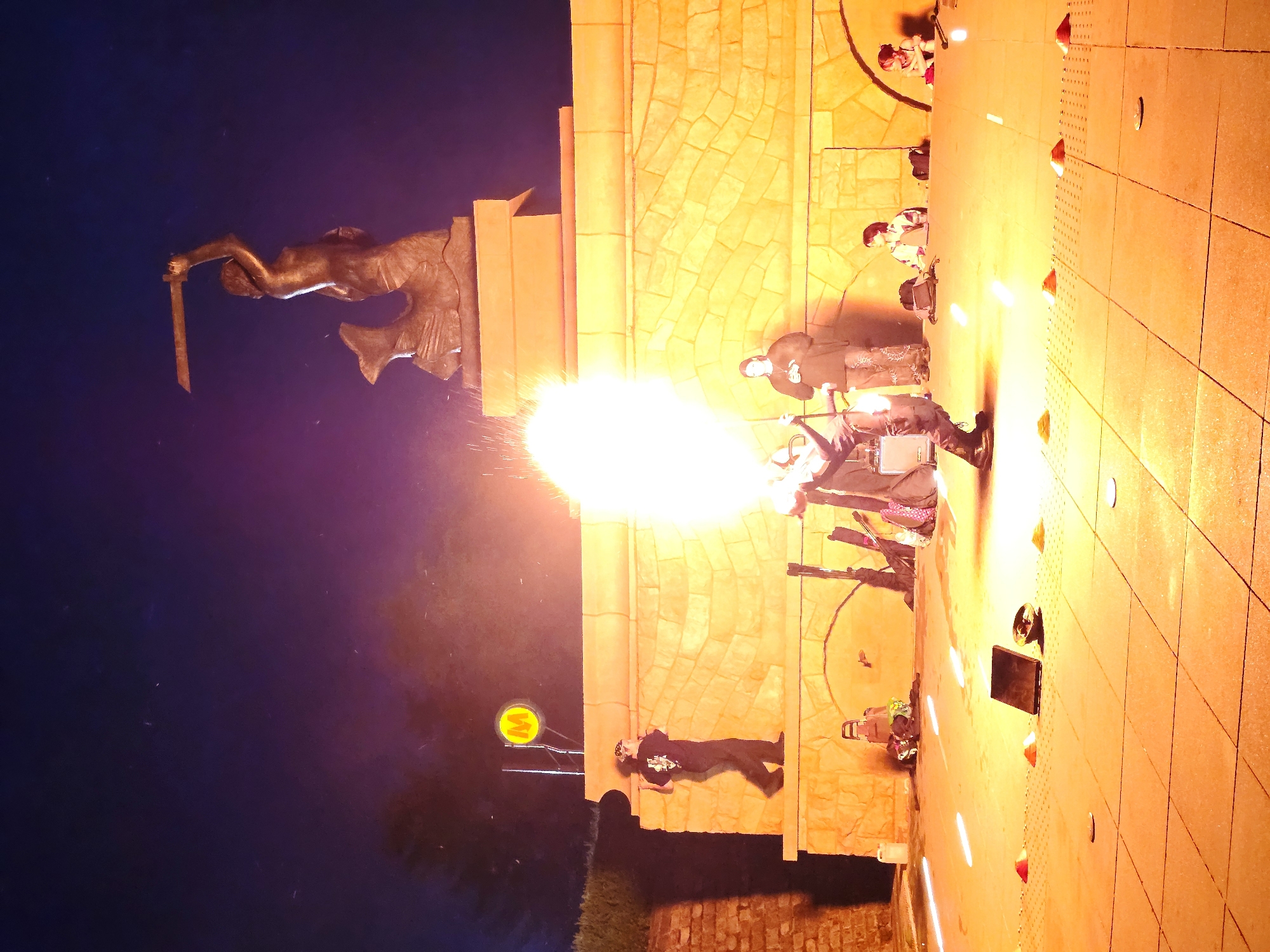 The image captures a lively night scene in Warsaw, Poland, likely near the Mermaid Monument, given its iconic silhouette against the night sky. A fire performer is the central focus, spewing a dramatic burst of flames that illuminates the surrounding area with an intense orange glow. The performer, dressed in dark clothing and a bandana, stands on a raised, stone-clad platform, actively engaged in the performance.
Behind the performer, the bronze statue of the Mermaid of Warsaw is visible, a symbol of the city, holding her sword aloft. The monument is situated on a raised base, also made of stone. To the left of the performance area, a circular yellow sign with a red "M" is mounted on a pole, suggesting proximity to a metro station.
Several spectators are gathered, observing the show. Some are seated on the ground in the foreground, while others stand further back near the platform. A few individuals are visible on the platform itself, seemingly part of the performance or assisting.
The ground in the foreground is paved with square tiles and features several small orange cones, possibly marking the performance area. The overall atmosphere is dynamic and festive, with the contrast between the dark night sky and the bright flames creating a visually striking spectacle. The lighting casts long shadows and highlights the textures of the stone structures and the performers.