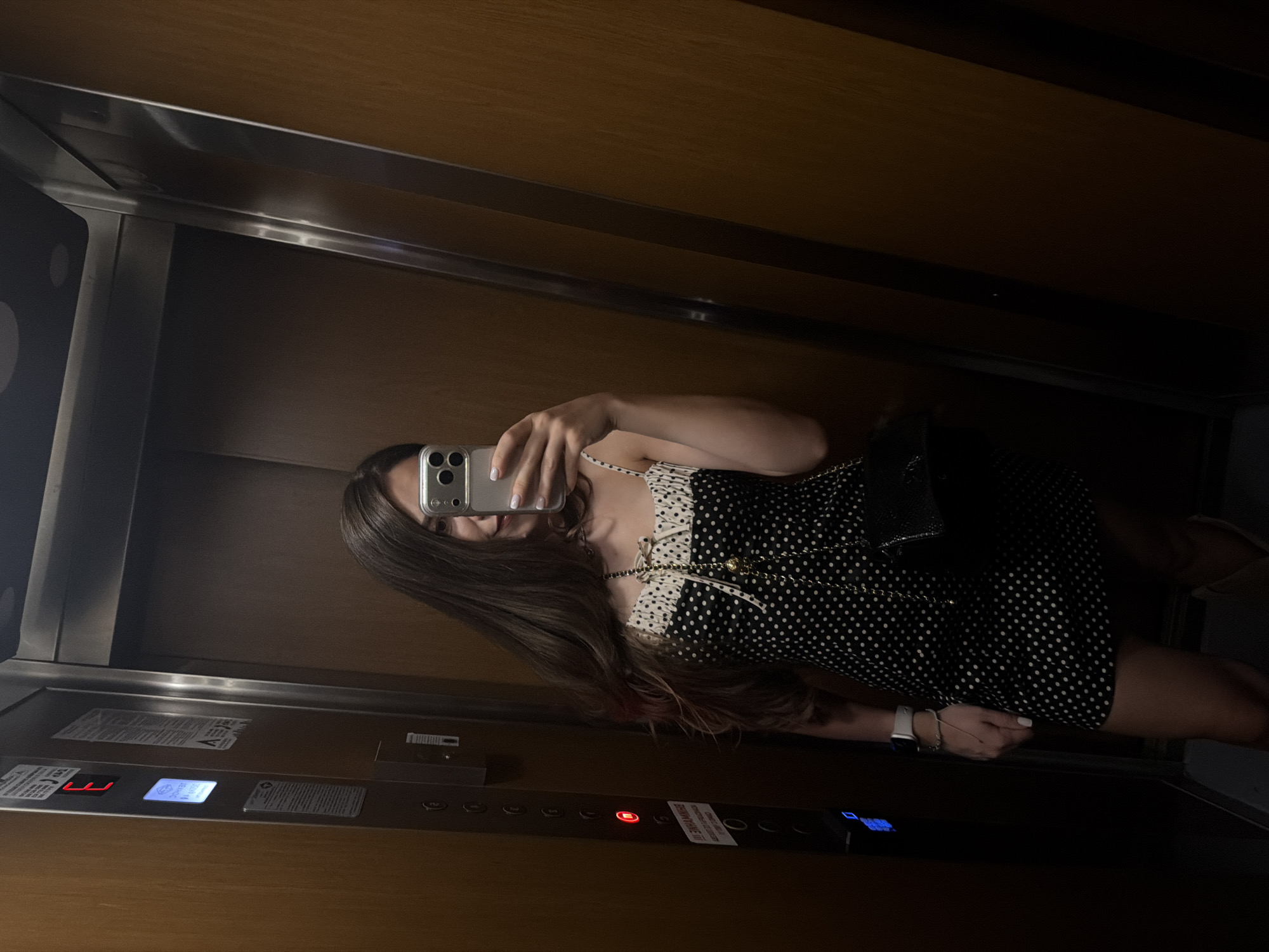 A young woman with long, dark brown hair is taking a mirror selfie in an elevator. She is wearing a black and white polka dot dress with a ruffled neckline and bows. She has a black handbag slung over her shoulder and is wearing a smartwatch. The elevator has wood-paneled walls and a panel of buttons on the left side, with one button illuminated red, indicating the current floor. The image appears to be taken in Razlog, Bulgaria, given the context of the location.