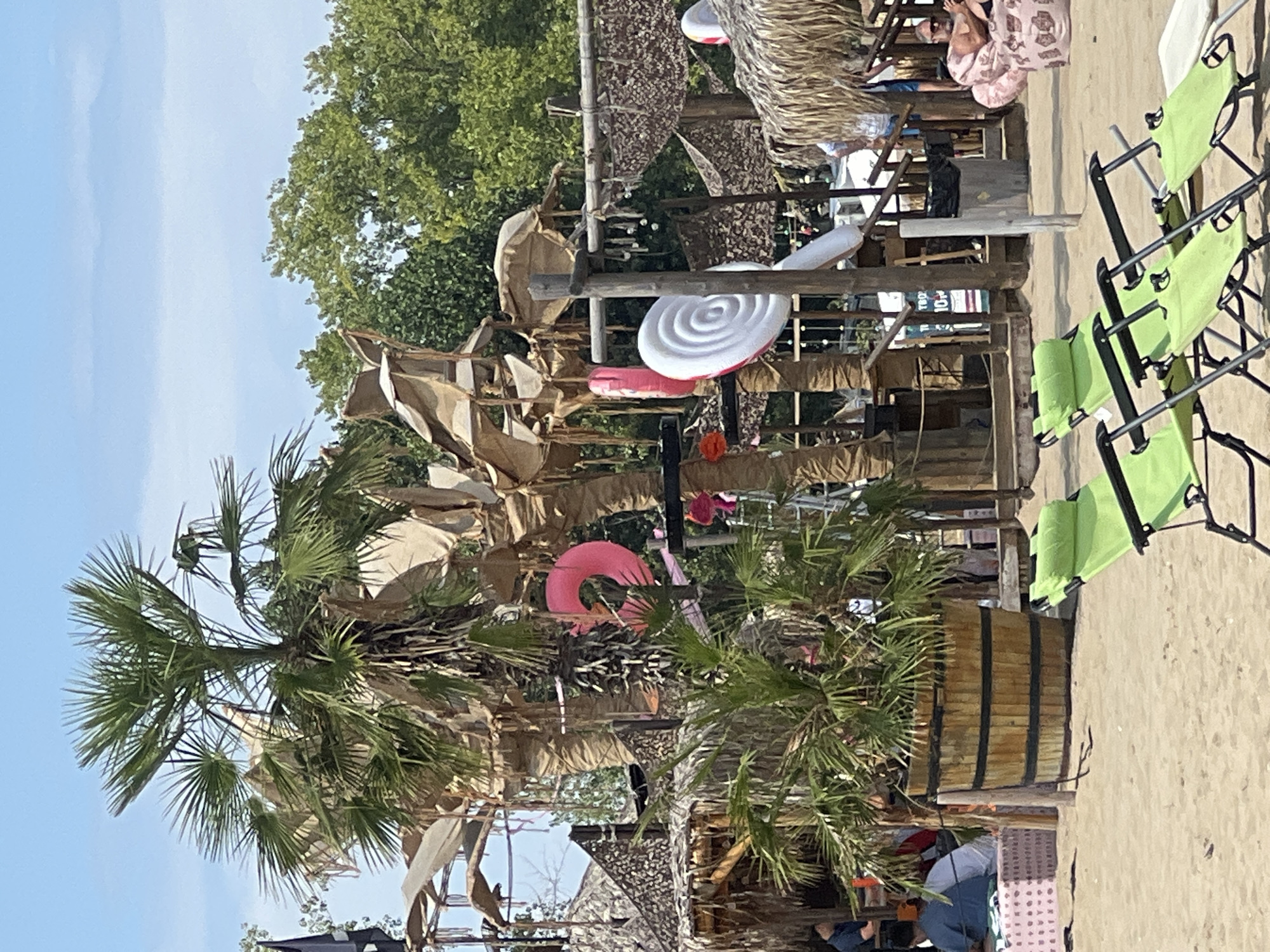 The image captures a sunny outdoor scene at a beachside establishment in Sozopol, Bulgaria. In the foreground, three lime-green lounge chairs are set up on a sandy beach, with the ocean implied but not directly visible. Behind the chairs, a large wooden planter holds a palm tree with dry, tattered fronds, possibly a stylistic choice for the venue. Further back, a rustic, open-air structure is visible, constructed with wood and thatched roofing, resembling a beach bar or lounge area. The structure is adorned with decorative inflatable objects, including a large pink ring float and a white spiral lollipop. Palm trees and lush green foliage form the backdrop. Several people are present in the background, some seated at tables and others standing, suggesting a lively atmosphere. The overall impression is a casual, tropical-themed resort or entertainment venue.