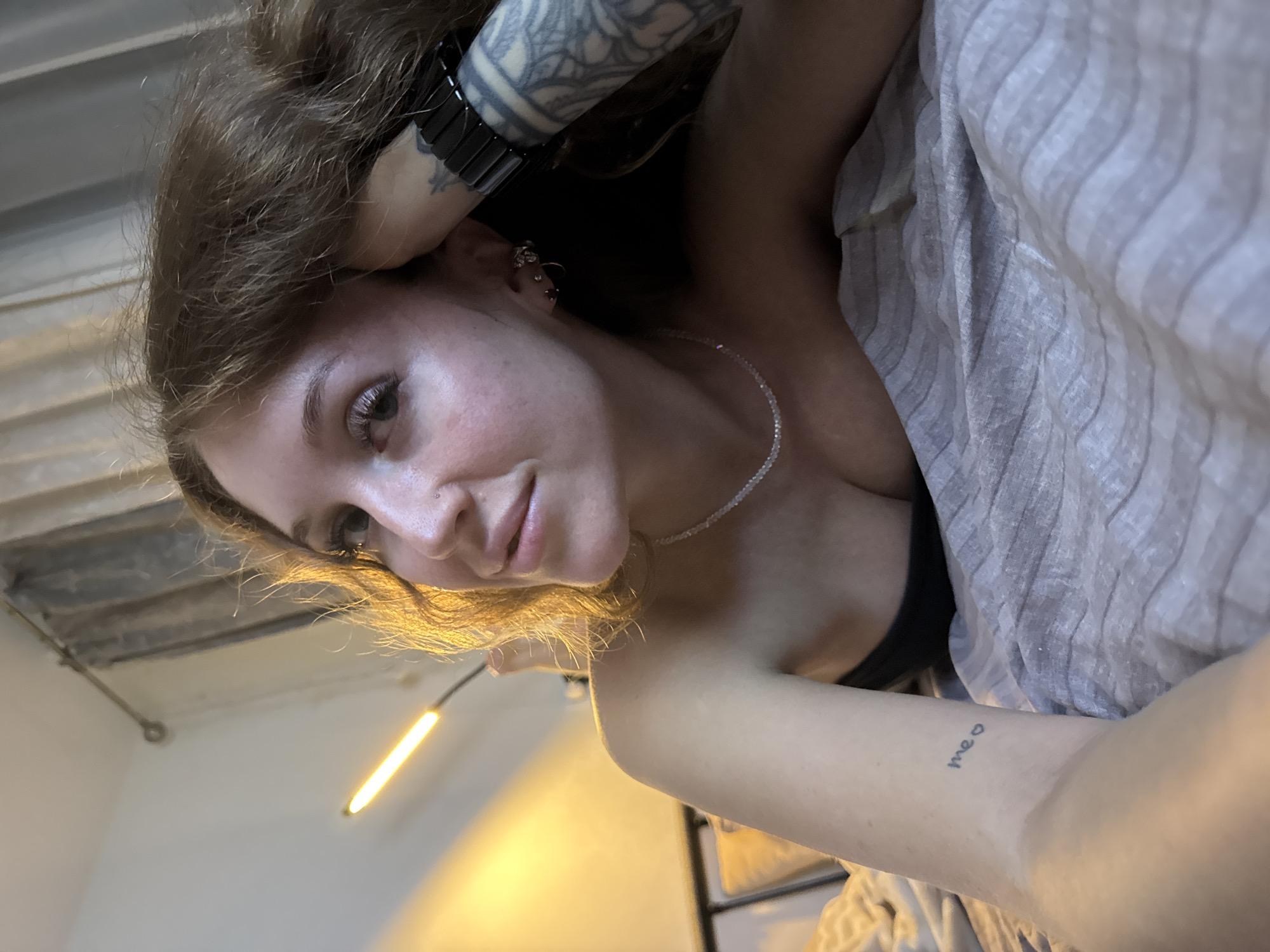 A woman with long, brown hair and visible tattoos on her arm and wrist lies on a bed covered with striped grey linens. She is wearing a strapless black top and a delicate necklace. Her head is propped up on her hand, and she is looking to her left. A bright lamp with a warm glow is positioned behind her, casting light on the wall and curtains. The scene appears to be indoors, likely a bedroom. There is no information or visual evidence to suggest this location is in Chaikovsky, Russia.