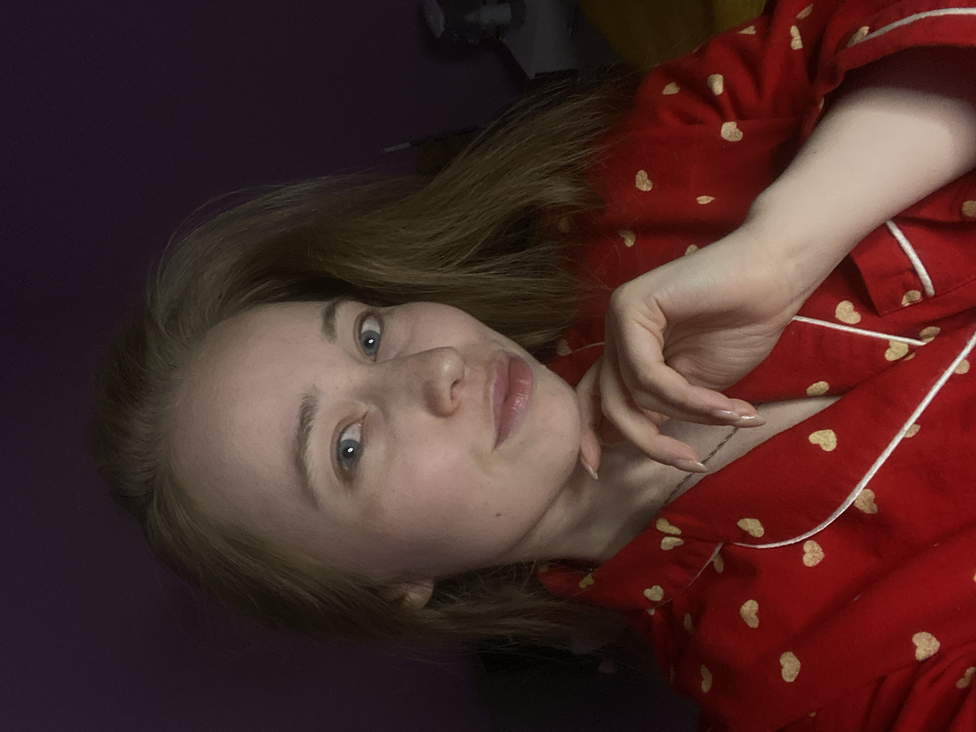 A light-skinned woman with fair hair and blue eyes is pictured in a close-up shot. She is wearing a red pajama top with small gold hearts and white trim, and her hand is positioned near her chin. The background is dark, and a partially visible object is in the upper-middle area. The image does not contain enough information to determine the location, so it is not possible to specify if it is in Saint Petersburg, Russia.