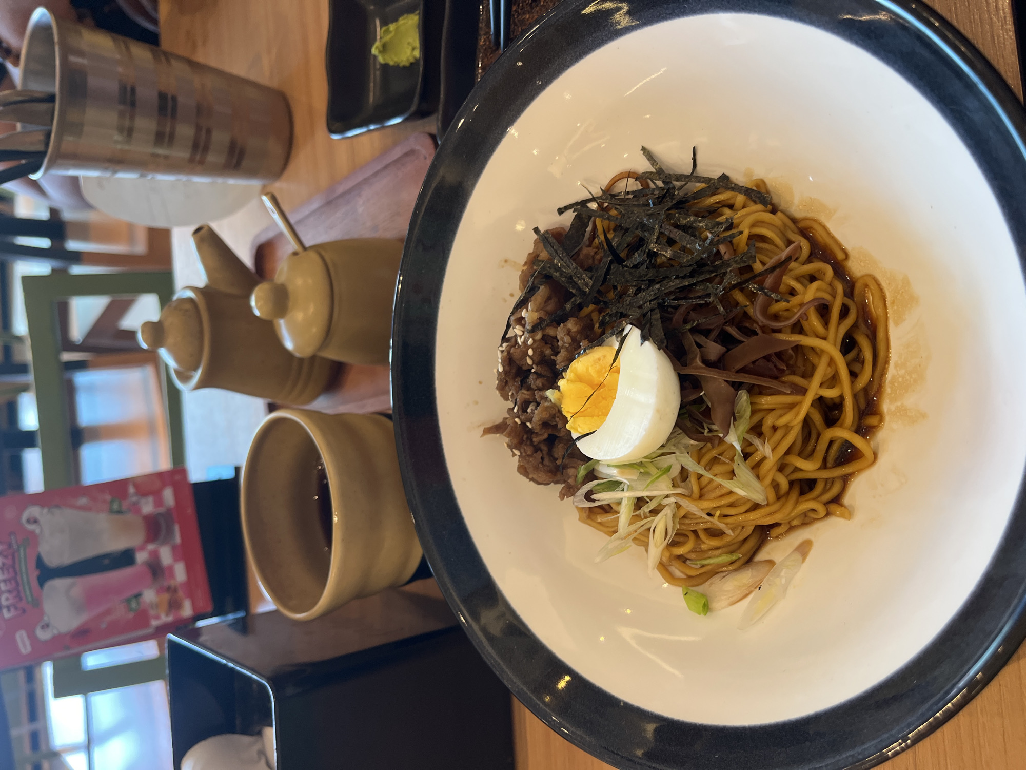 The image shows a bowl of what appears to be Japanese-style dry ramen. The noodles are topped with shredded seaweed, cooked meat (possibly beef), half of a boiled egg, and sliced green onions. The bowl has a white interior and a dark gray or black rim. The bowl sits on a wooden table. To the left of the bowl are a set of ceramic jars (likely for sauces) and a cup. Further to the left, there's a container holding chopsticks and a pamphlet, which is partially visible. In the background, there's a glimpse of the interior of a restaurant, with tables, chairs, and a window.
While the description request included the city of Lhokseumawe, Indonesia, there is no identifying information in the image that definitively places the scene in that location. Therefore, the location cannot be confirmed from the provided image.