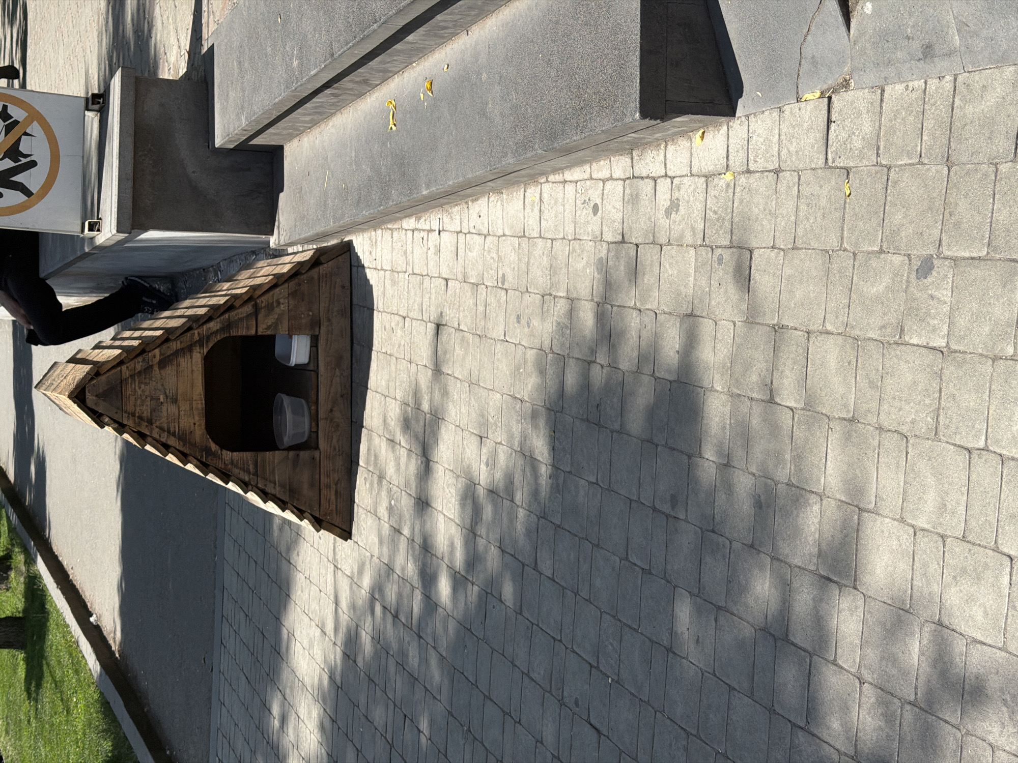 The image shows a wooden dog house shaped like a triangle with a peaked roof. Inside the dog house, there are two plastic bowls, one of which appears to contain water. The dog house is situated on a paved area with square gray pavers. To the right of the dog house are concrete steps, and a person's legs are visible, wearing black pants and dark shoes, sitting on a concrete ledge. A sign with a "no dogs allowed" symbol is also present on the ledge. The scene appears to be outdoors, possibly in a park or public space. The lighting suggests it is daytime, with shadows cast on the paved ground. The location is identified as Yerevan, Armenia.