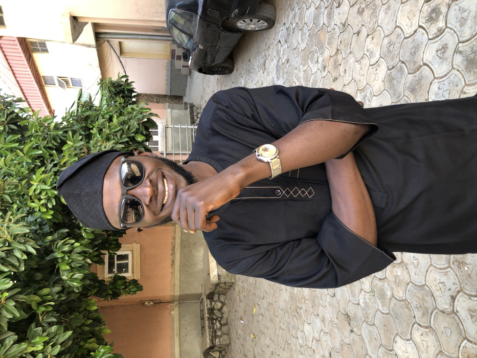 A dark-skinned man is captured in a vertical, medium-shot photograph, standing outdoors in what appears to be a paved courtyard in Lankan, Nigeria. He is wearing a black traditional outfit, possibly a dashiki or caftan, featuring subtle light-colored embroidery in a diamond pattern on the chest and cuffs. A matching black cap covers his head, and he sports dark sunglasses, which reflect the bright surroundings. A prominent gold-toned watch with a metal strap is visible on his left wrist. The man has a beard and is smiling broadly at the camera, showing his teeth, conveying a cheerful and friendly demeanor. His arms are crossed in front of him.
The background features dense green foliage from bushes or trees on the left side of the frame. Parts of a light-colored building with visible windows are situated behind the man and the greenery. To the far right and in the distance, a black car is parked. The ground he stands on is paved with distinctive interlocking concrete blocks forming a wavy or scalloped pattern. The lighting suggests a bright, sunny daytime setting, likely mid-day or early afternoon, indicated by the strong illumination and visible shadows. There is no visible text in the image.