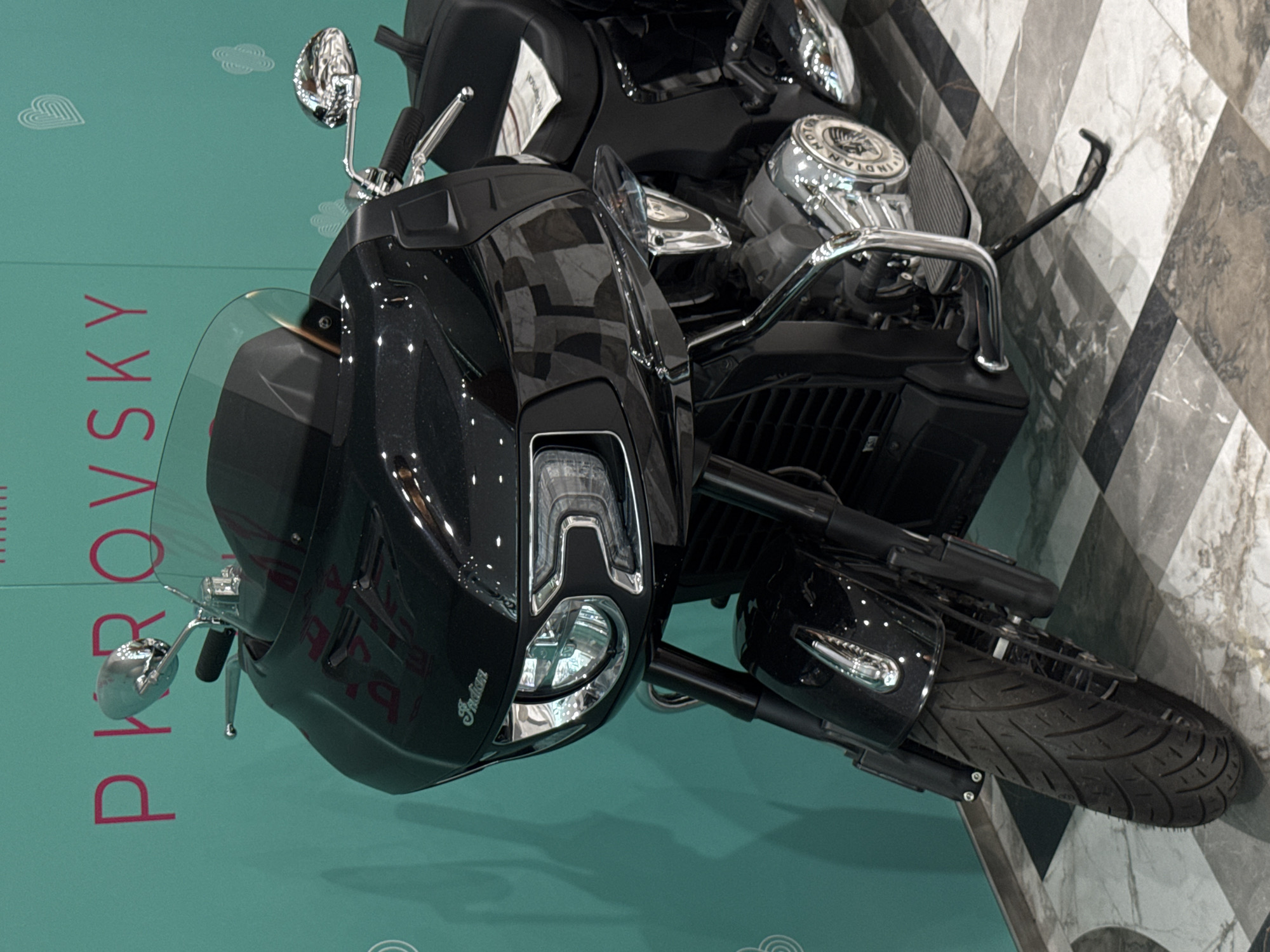 A black Indian motorcycle is parked indoors, possibly in a showroom or at an event. The motorcycle has a large front fairing, a windshield, and chrome accents. The brand "Indian" is visible on the motorcycle's body. In the background, there is a teal-colored wall with pink lettering that reads "PIROVSKY" and some decorative heart shapes. The floor is tiled with a checkered pattern of gray and black marble-like tiles. The lighting suggests an indoor environment, likely with artificial illumination. The image does not provide any explicit clues about the specific location within Yekaterinburg, Russia, other than the presence of the brand name "PIROVSKY" on the wall, which could be a store, event, or company name.