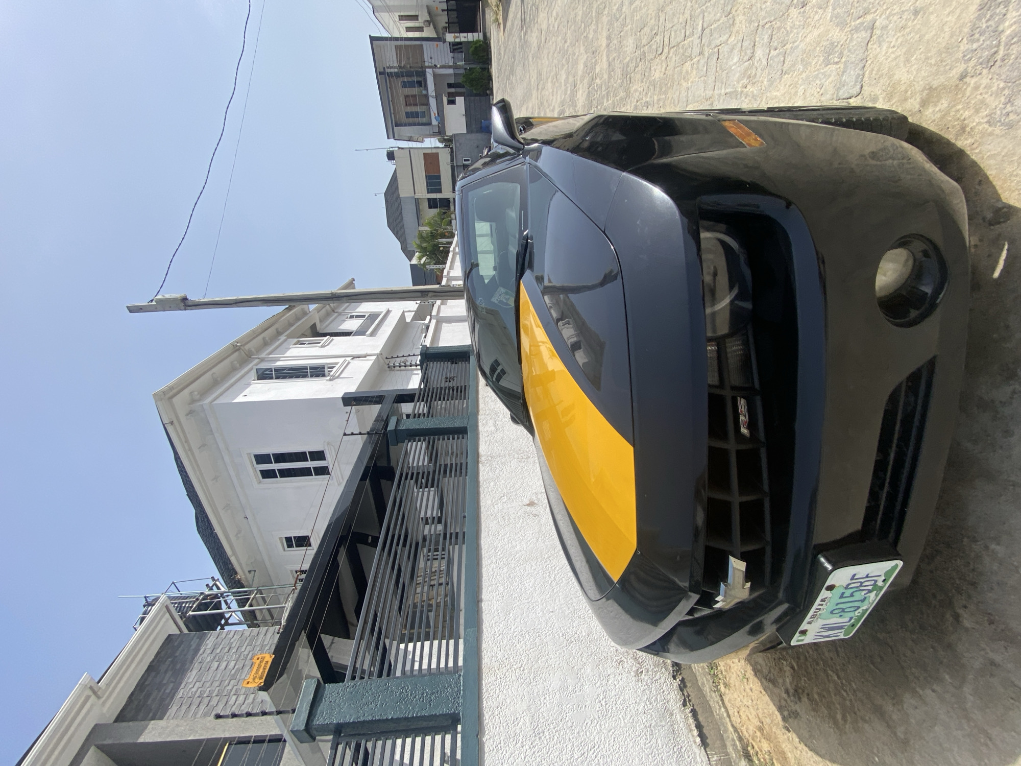 A black and yellow Chevrolet Camaro is parked on a paved surface next to a white wall and black metal fence. A multi-story white building with black-framed windows is visible in the background. The car has a license plate that reads "KWL-815BE" and "ABUJA". The sky is blue with sparse clouds. The location is likely in Lagos, Nigeria, based on the license plate style.