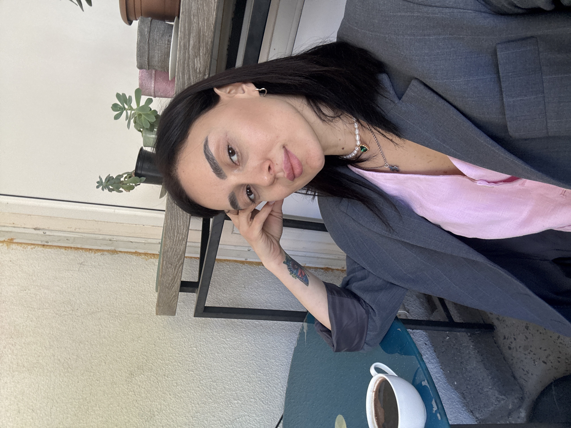 A woman with dark hair is seated outdoors at a teal blue table. She is wearing a gray blazer over a pink top, and a pearl necklace. A white coffee cup sits on the table in front of her. She is resting her head in her hand, and a butterfly tattoo is visible on her forearm. The background includes plants in pots on a wooden shelf, and a white wall. The location is possibly in Tbilisi, Georgia, suggested by the style of outdoor seating and the architectural elements.