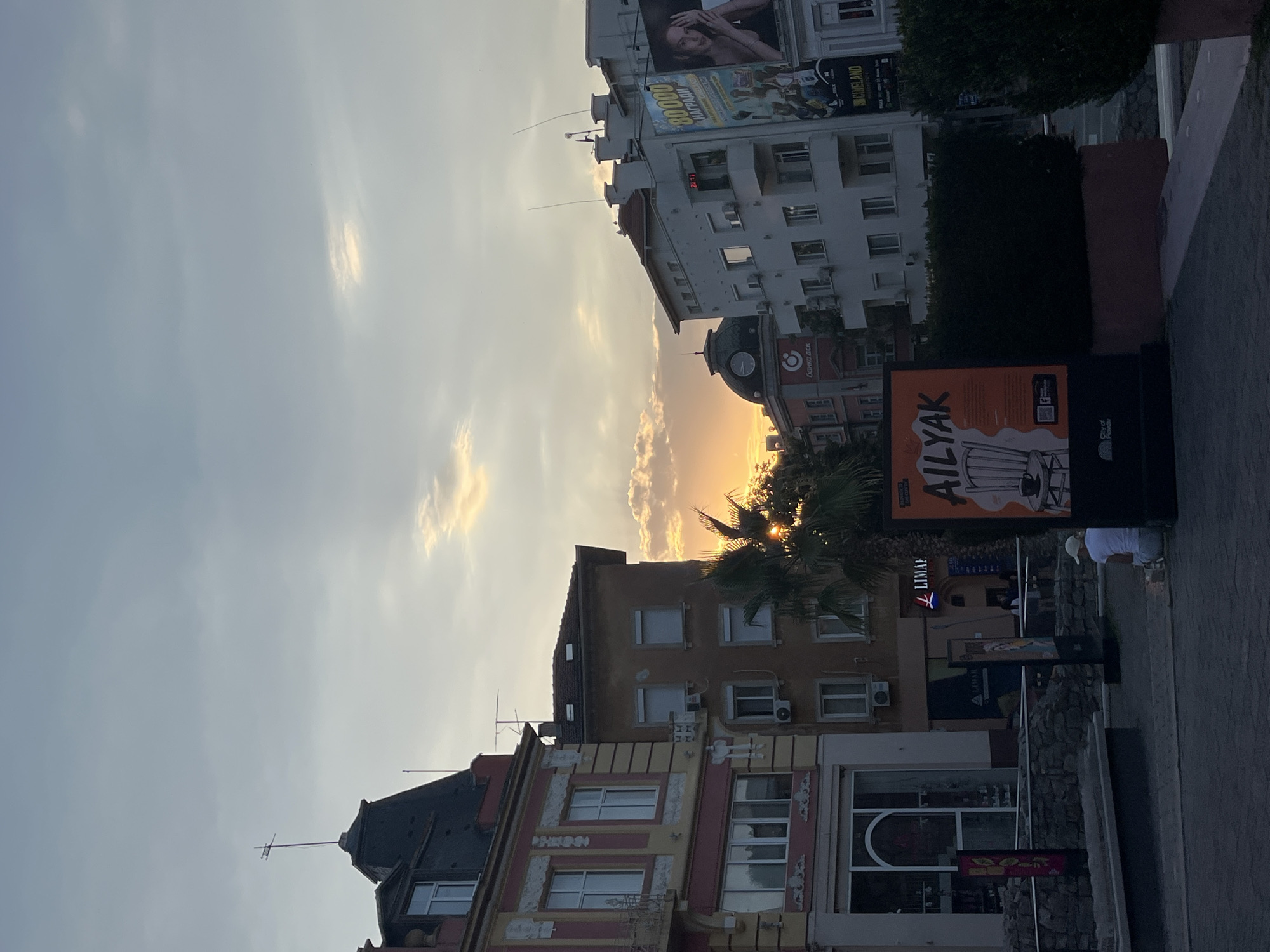 The image depicts a street scene in Plovdiv, Bulgaria, during what appears to be sunset. In the foreground, a person wearing a white cap and a white t-shirt is sitting on the ground, facing away from the camera. To the left, a large advertisement board features the text "AILYAK" in orange and white lettering, with an illustration of a chair and a cup.
The middle ground shows several buildings with architectural details characteristic of European cities. The building on the far left has a red and yellow facade with decorative elements and large windows. Adjacent to it is a brown building with numerous windows and air conditioning units. A palm tree is visible between these buildings, with its fronds partially obscuring the setting sun, which is visible as a bright orange glow behind the trees and buildings.
In the background, more buildings are visible, including a white building with a clock tower on its facade. To the right, a billboard advertises "80000 НАГРАДИ" (80000 Awards) and features an image of a woman. The sky is overcast with soft clouds, and the overall lighting suggests the golden hour. The street appears to be paved, and a low stone wall is present in the middle ground. The scene conveys a sense of urban tranquility as day transitions into evening.