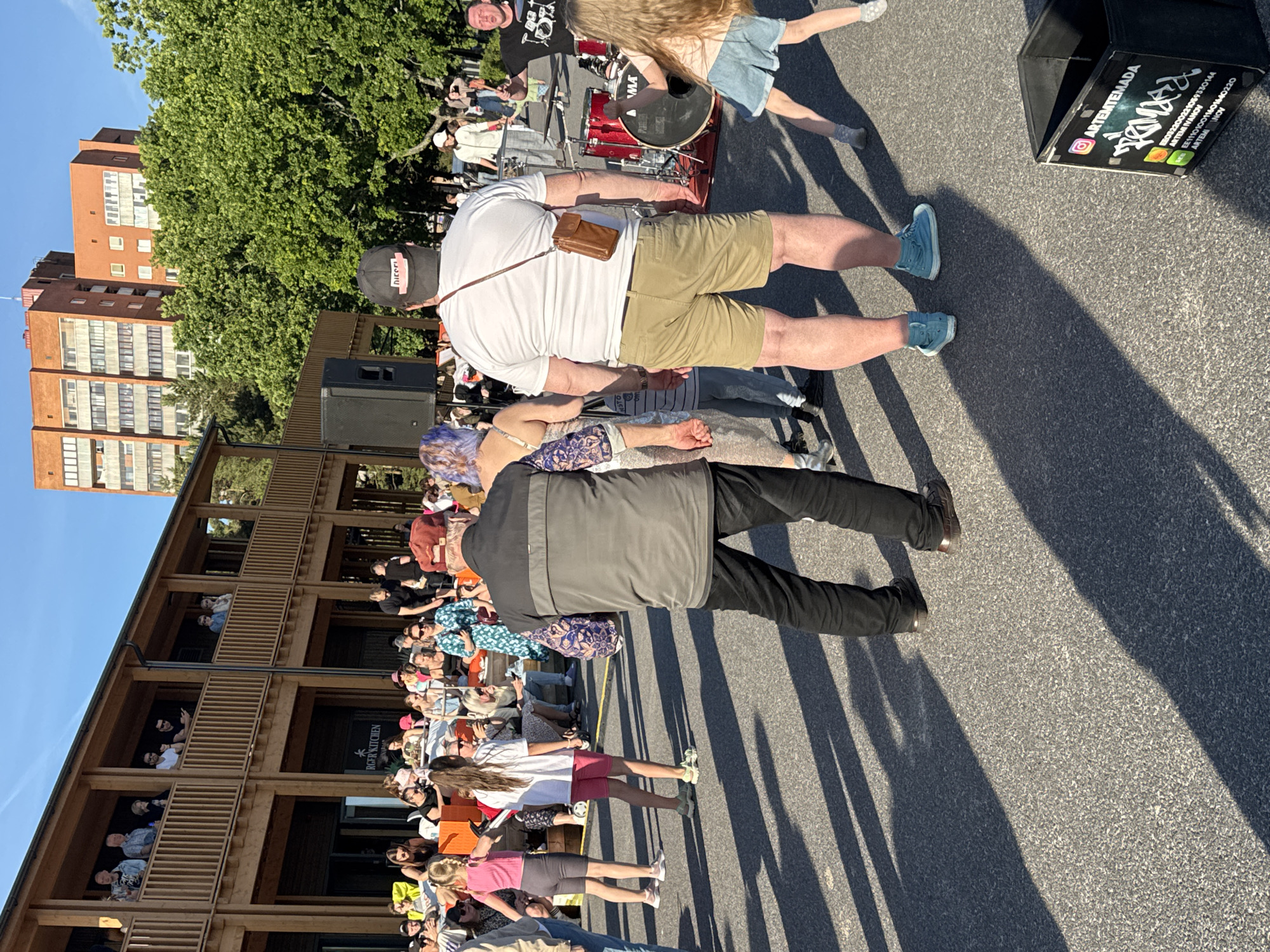 In Tallinn, Estonia, an outdoor gathering is taking place near a building with a wooden facade and balconies. The sun casts long shadows across the asphalt ground. In the foreground, a man in a grey jacket and black pants is facing away from the camera, while another man in a white t-shirt and khaki shorts is also facing away. A young girl in a light blue denim skirt and pink top is dancing. In the background, a crowd of people are seated and standing, some near a stage with musical instruments, including drums. A tall brick building is visible beyond the trees.