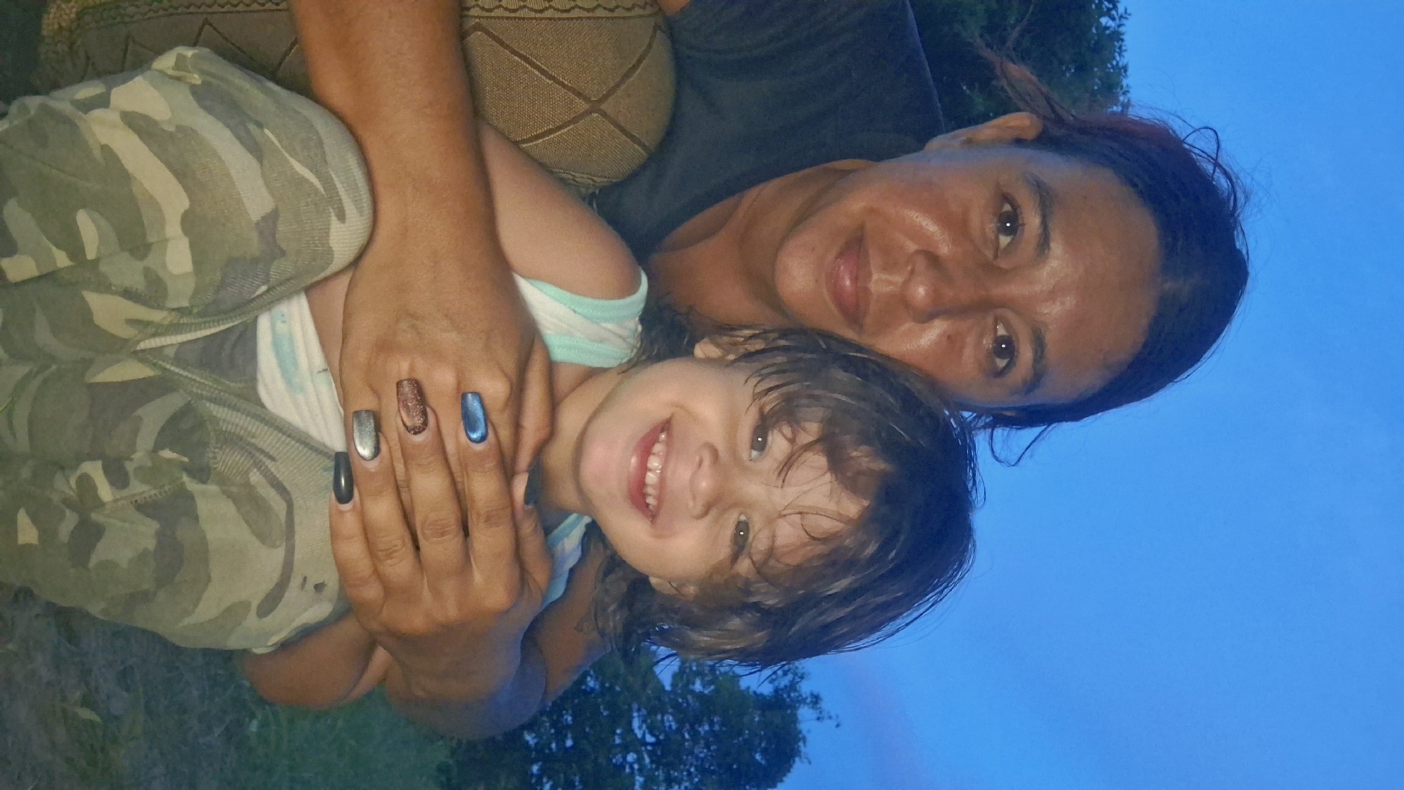 In the city of Vallecito, Venezuela, a woman and a young child are captured in a photograph against a twilight sky. The woman, with dark hair and tanned skin, is positioned behind and to the left of the child, her arms wrapped around the child's torso. She is wearing a dark t-shirt. The child, facing the camera with a wide smile, has dark, tousled hair and is wearing a white tank top with thin blue stripes. The child's hands are resting on the woman's arms, and the woman's fingernails are painted in a mix of blue, silver, and glittery brown. The child is wearing camouflage-patterned pants. To the left of the woman, a portion of a textured, possibly upholstered, surface is visible. The background consists of dark trees and the deep blue of the evening sky.