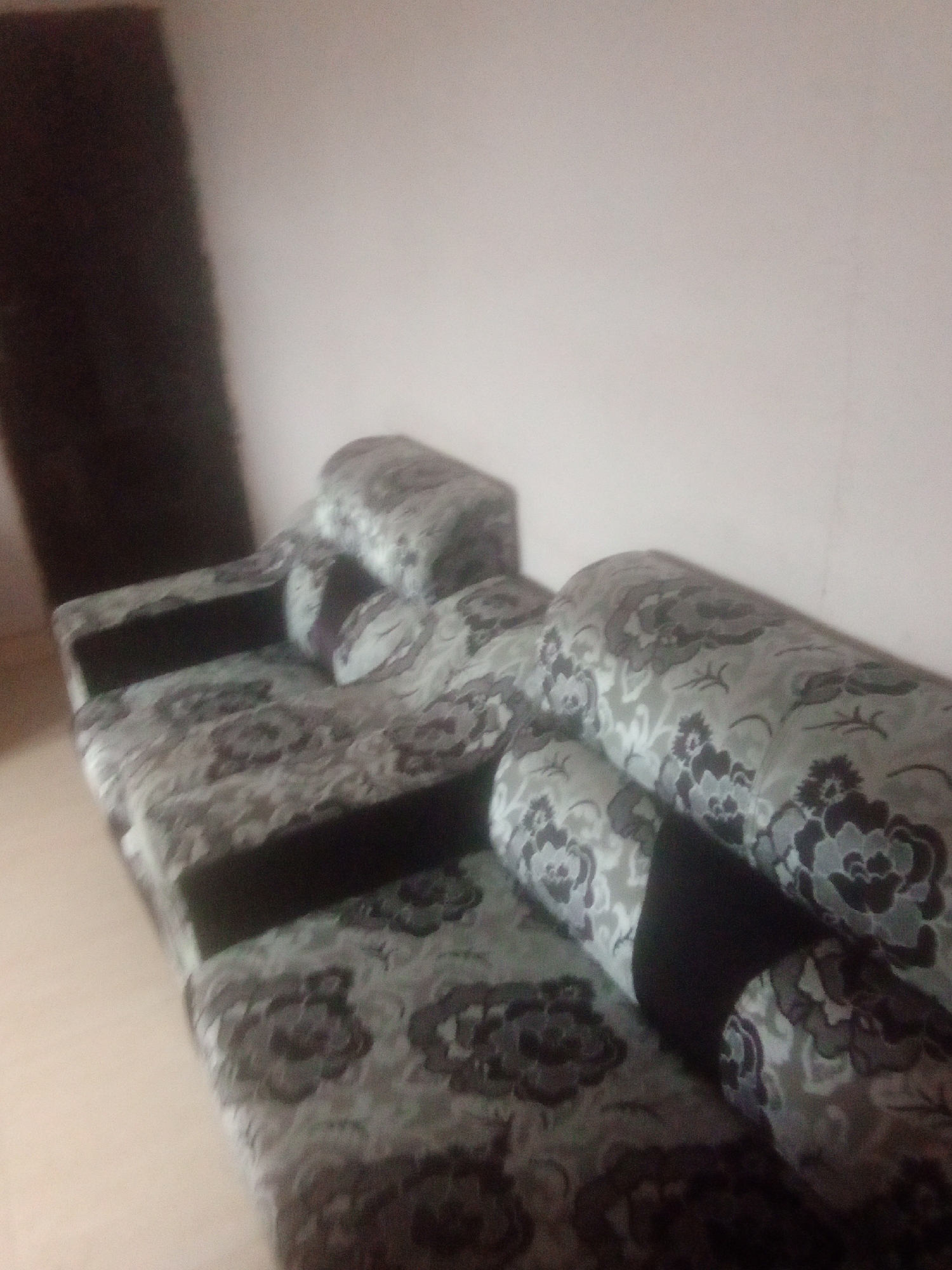 The image shows a large sectional sofa with a floral pattern. The sofa is upholstered in a fabric with a base color of light gray and a pattern of dark gray and black flowers. The sofa appears to be in a living room, with a plain, light-colored wall in the background. To the left of the sofa, a dark, vertical object is visible, possibly a piece of furniture or a doorway. The lighting in the image is dim, suggesting it might be evening or indoors with artificial light. The scene is static, with no people or activity taking place. The focus is on the furniture, which fills most of the frame. The overall impression is that of a simple, domestic setting. There is no visible text in the image. The location context of Jalingo, Nigeria does not provide any specific visual cues within the image itself.