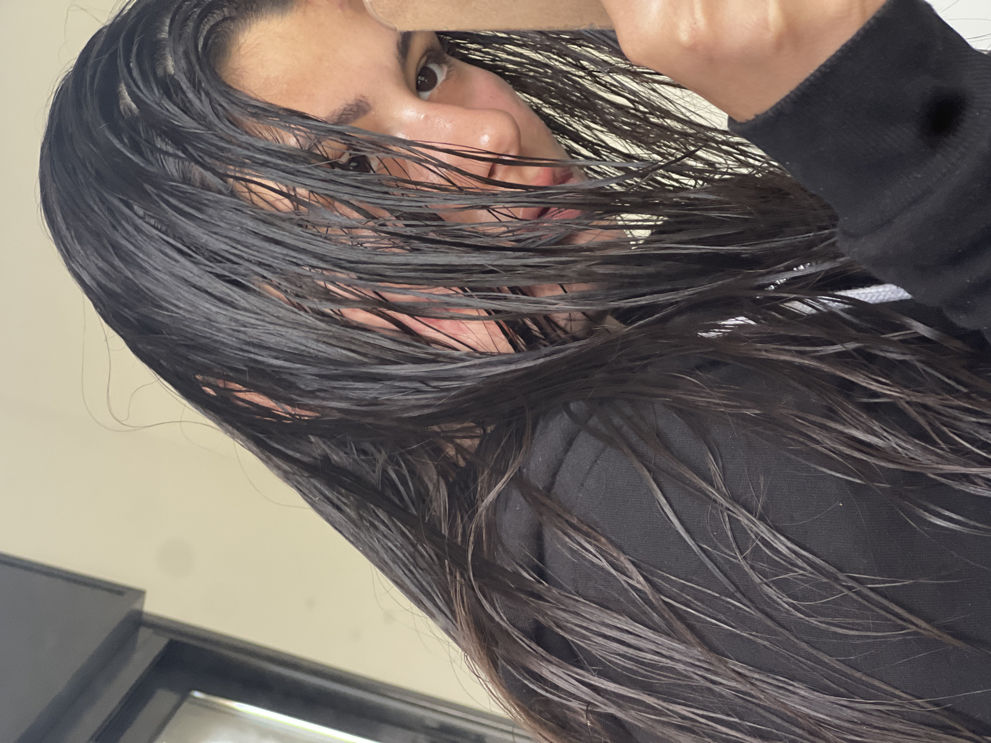 A person with long, dark, wet hair is seen from a low angle, obscuring much of their face. Their skin appears oily or wet. They are wearing a black hooded sweatshirt. The background includes a light-colored wall and what appears to be a window frame or part of a door. There is no information to place this image in Kiryat Bialik, Israel.