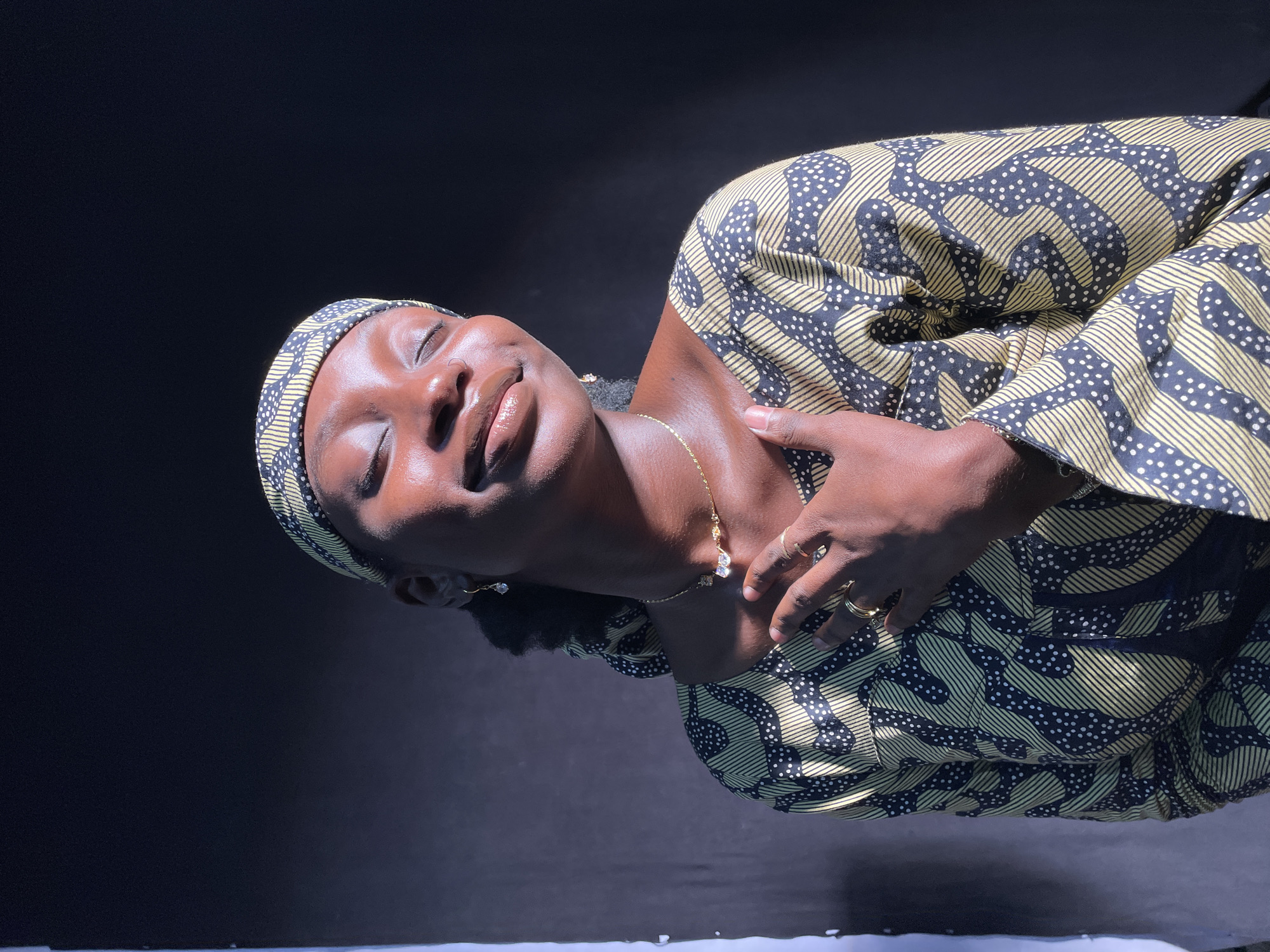 A portrait of a person, likely female, is set against a dark, featureless background, captured in Jos, Nigeria. The subject, with dark skin, is shown from the chest up, with their head tilted gently to the right and eyes closed, conveying a serene and contemplative expression. Their lips are slightly parted, and a strong, bright light source illuminates the left side of their face and upper body, creating highlights on their skin.
The person wears a headwrap and a matching long-sleeved garment, possibly a dress or top, both made from a distinctive patterned fabric. The design consists of black with light yellow or gold wavy lines and scattered white dots. Adornments include a slender gold chain necklace with a small, clear-gemmed pendant (appearing butterfly-shaped), a delicate nose ring on their left nostril, and a small earring in their left ear. Several gold rings are visible on the fingers of their left hand, which is gently placed on their upper chest or collarbone area. No text is visible in the image.