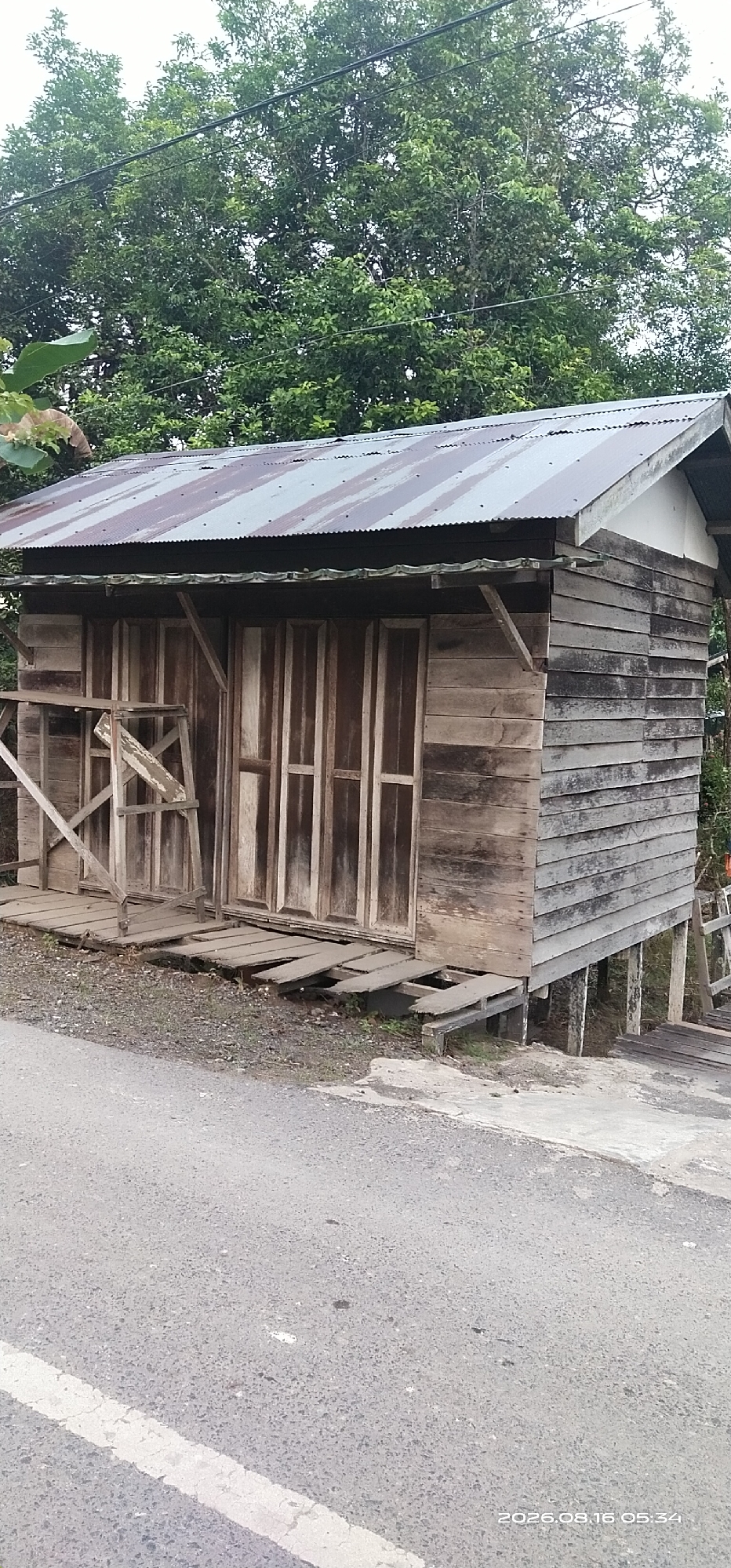 The image shows a dilapidated wooden shack on stilts next to a paved road. The shack has a corrugated metal roof and wooden plank walls. The front of the shack has large wooden double doors. A wooden walkway leads to the shack, and some of the planks are broken and missing. The shack is surrounded by lush green trees. A white line is painted on the road in the foreground. The timestamp in the lower right corner indicates the image was taken on August 16, 2026, at 05:34. The location is identified as Muka Haji, Indonesia.