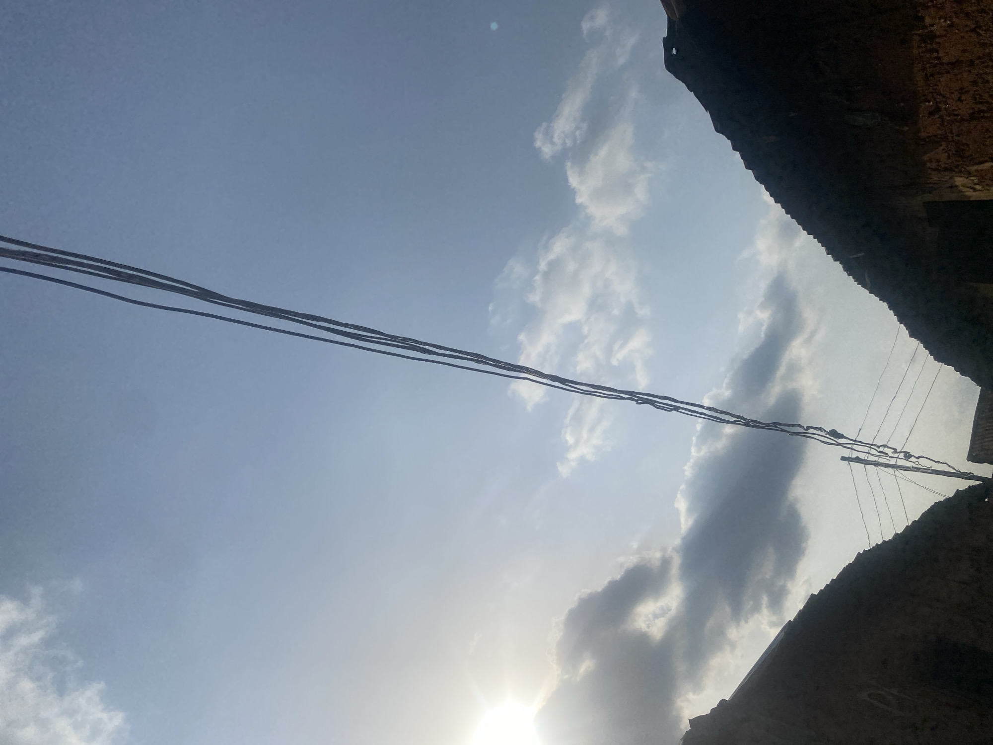 The image is taken looking upwards, capturing a view of the sky partially obscured by buildings and utility lines. The sky is predominantly blue with scattered white and grey clouds. The sun is visible near the bottom, creating a bright glare. Utility lines, including some thicker cables, stretch across the frame from left to right. Dark, textured structures that appear to be the roofs or walls of buildings are visible at the top right and bottom right corners of the image. The location is in the city of Unknown, Unknown.