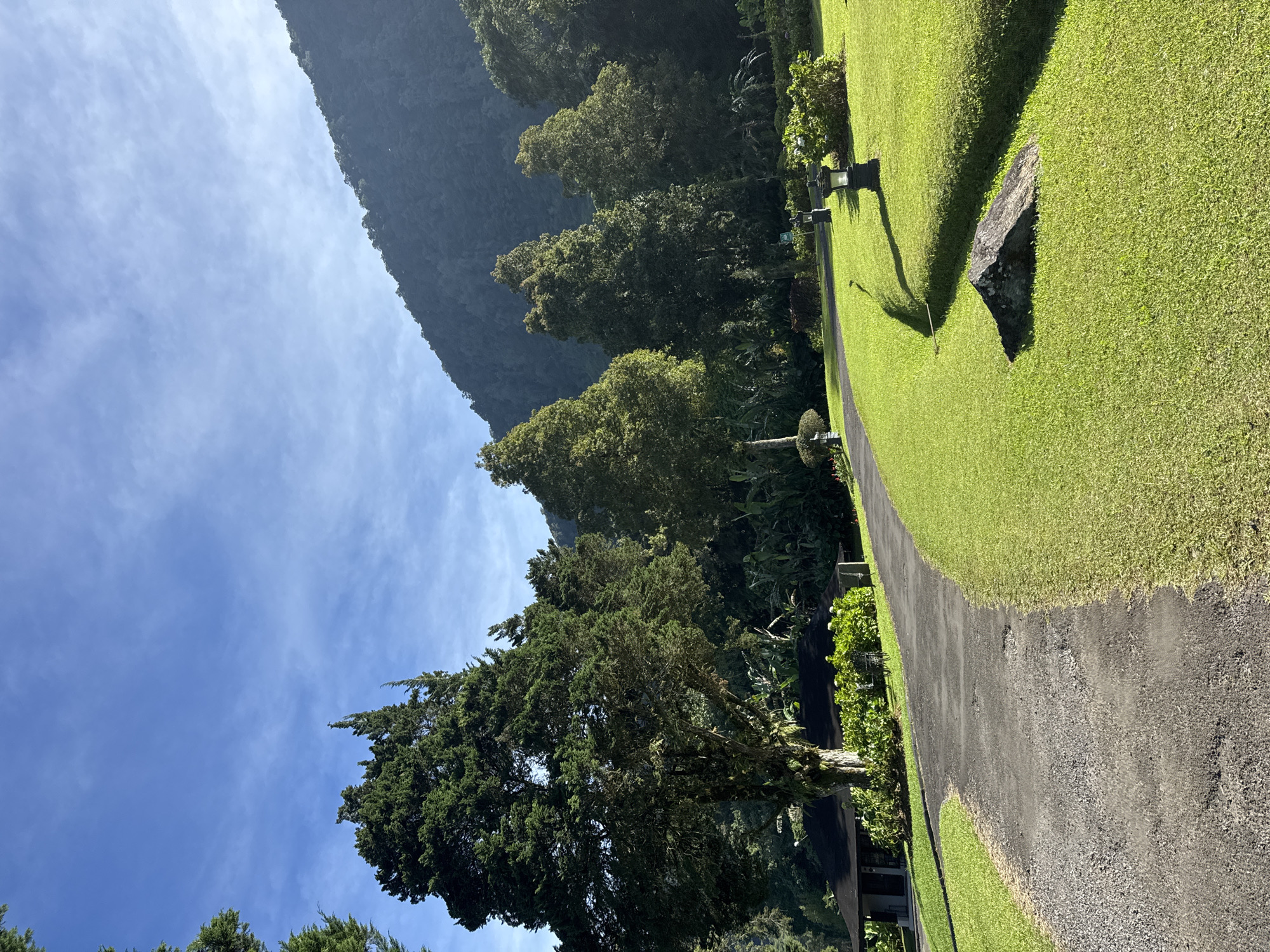 The image shows a landscape with a paved pathway running diagonally from the lower right to the upper left. Green grass covers the areas on either side of the path. To the left of the path, there is a line of trees of varying heights. In the background, a mountain range rises up to the left. The sky is blue with scattered clouds. A stone is positioned on the right of the pathway. The location is in the city of Unknown, Unknown.