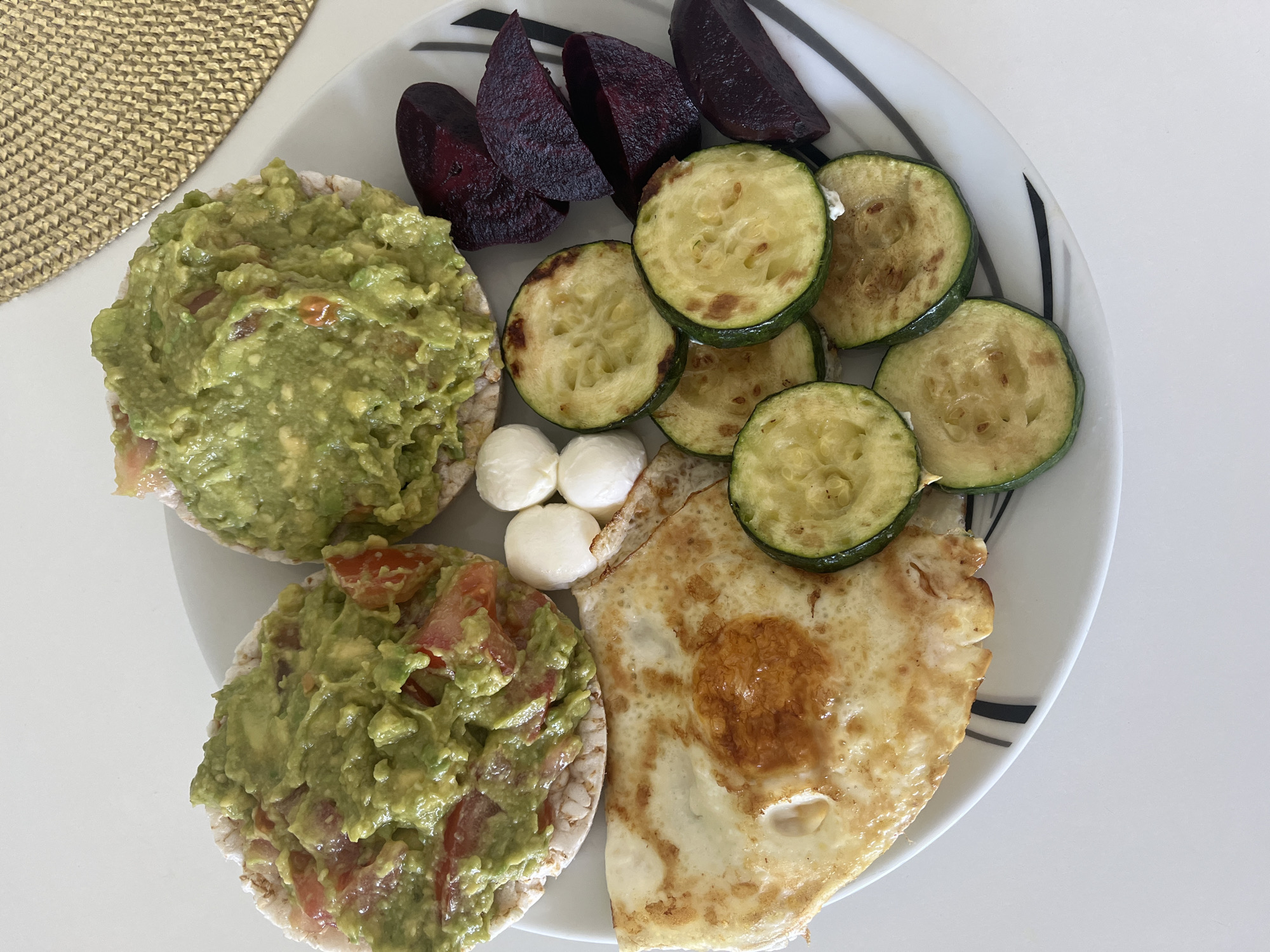 On a white plate with black stripes sits a meal consisting of various foods. Two rice cakes are each topped with a generous serving of guacamole, the bottom one having visible pieces of tomato. Beside the rice cakes, there are three small, round, white mozzarella balls. Adjacent to the mozzarella, a fried egg is visible. Next to the egg, a row of grilled zucchini slices are arranged. In the top right corner, four pieces of cooked beet are placed. The plate is sitting on a white table, and a woven placemat is visible in the upper left corner of the frame. The image appears to be taken in a bright, well-lit environment. Location details are not specified.