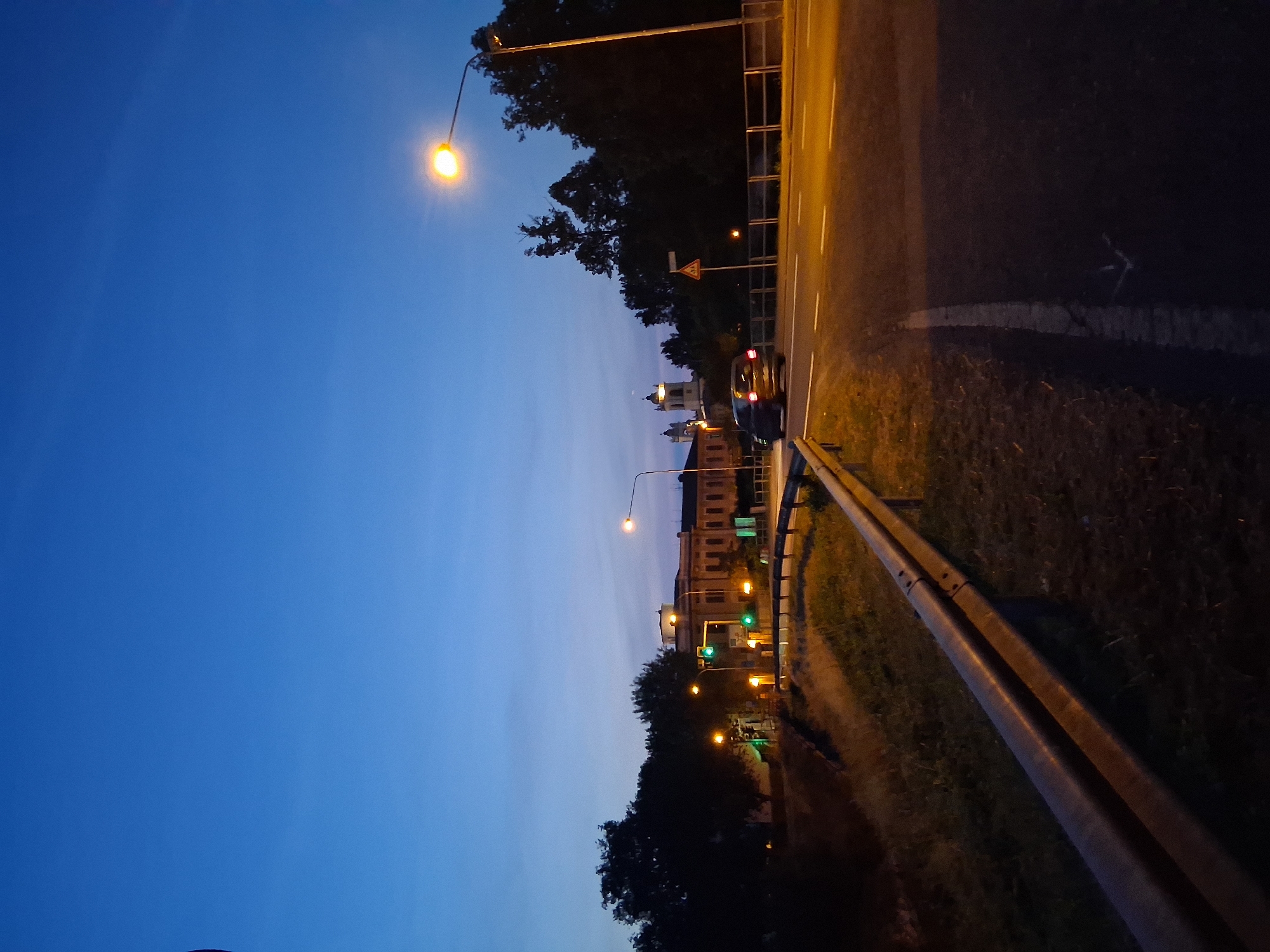 This image captures a street scene at dusk or dawn in Colorno, Italy, with the Ducal Palace prominently visible in the background. The sky is a clear, deep blue, transitioning from lighter shades near the horizon to darker overhead, with a faint crescent moon visible above the palace's bell towers.
In the foreground, a two-lane asphalt road with distinct white and yellow markings curves to the right. A metal guardrail runs along the left edge of the road, separating it from a grassy embankment. A single dark-colored car, possibly a hatchback or SUV, is traveling away from the viewer on the right lane, its red taillights illuminated.
Multiple artificial light sources define the scene. A tall streetlight on the far right illuminates the road with a warm orange glow. Additional streetlights are visible further down the road on the left. Green traffic lights are active on the left side of the road, indicating a cleared intersection or turn. A yellow triangular road sign, depicting a pedestrian crossing, stands on the right side of the road behind the car.
Dense, dark trees line both sides of the road in the mid-ground. Behind the trees on the left, a multi-story building with a light-colored facade and multiple windows is visible. To the right and behind this building, the distinctive twin bell towers and part of the main structure of the Ducal Palace of Colorno rise above the tree line, softly illuminated by distant lights. No people are discernible in the scene.