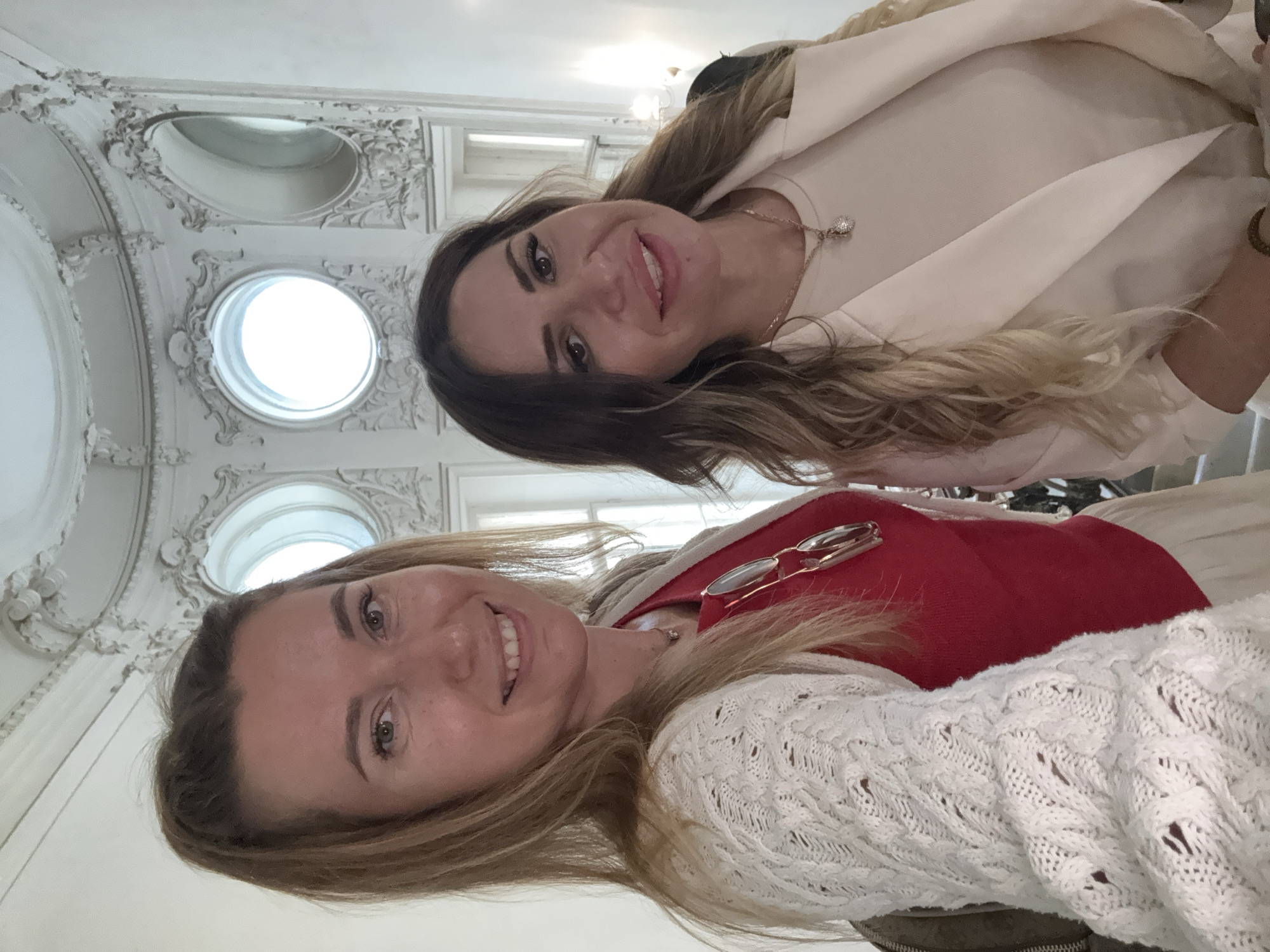 Two women are taking a selfie in a grand, ornate hallway with ornate architectural details. The woman on the left has blonde hair pulled into a ponytail and is wearing a red top and a white knitted cardigan. The woman on the right has long, wavy brown hair and is wearing a light-colored outfit. They are both smiling at the camera. The background features decorative molding, circular windows, and what appears to be a grand staircase.