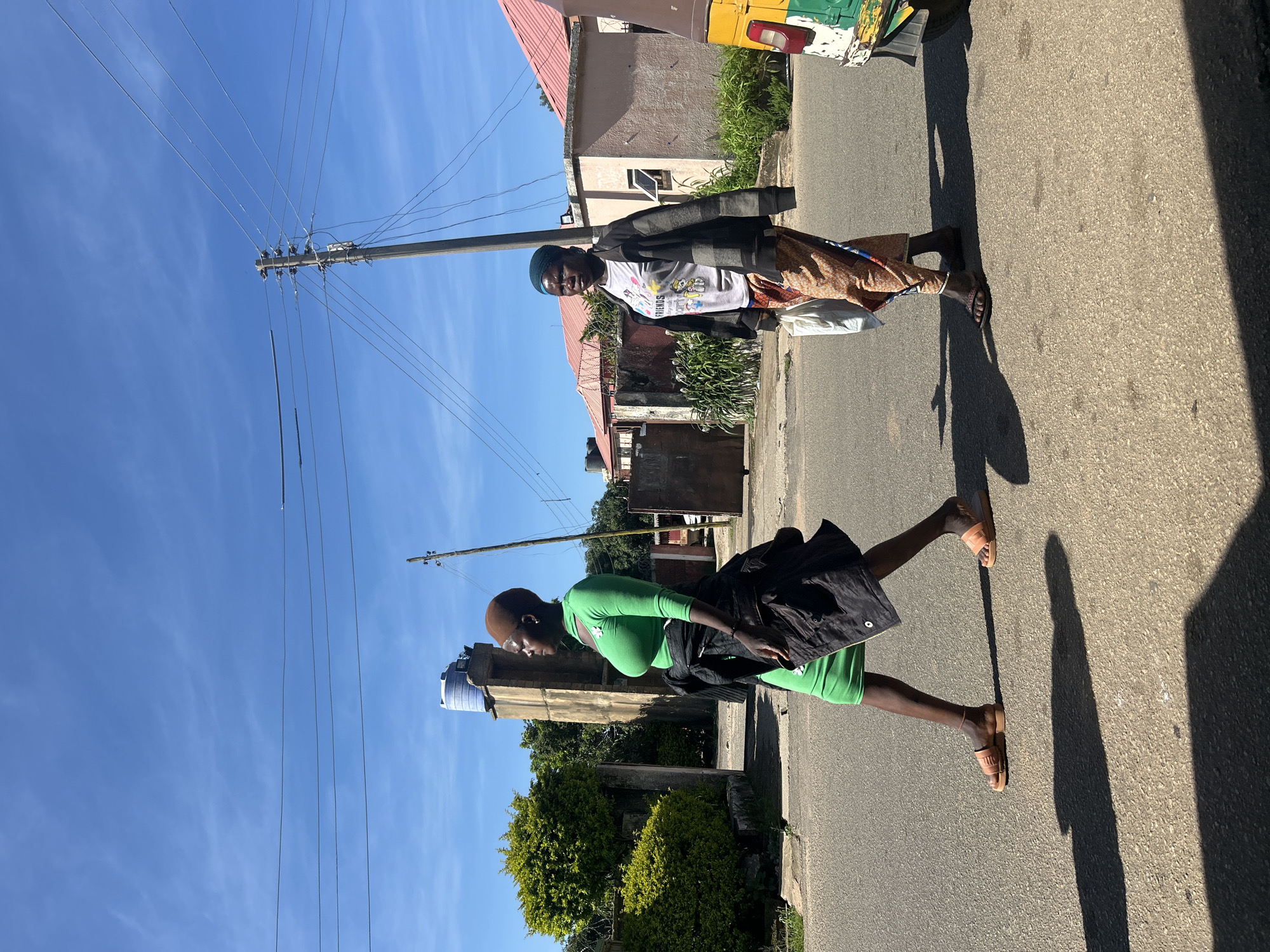 The image depicts an outdoor scene in Jos, Nigeria. Two women are walking on a paved road. The woman on the left is wearing a form-fitting green dress, a dark jacket tied around her waist, and light brown sandals. She has a brown headscarf and is looking to her right. The woman on the right is wearing a light-colored t-shirt with graphics, a dark jacket, and patterned trousers. She has a blue headscarf and is carrying a white plastic bag in her left hand. She is looking forward. Behind the women, there are buildings with pink roofs, a water tank on a tower, a utility pole with wires, and lush green bushes. A yellow and green vehicle, possibly a tuk-tuk, is partially visible on the right side of the frame. The sky is clear and blue with some faint clouds. The sun is casting long shadows on the road, indicating it is likely late afternoon.