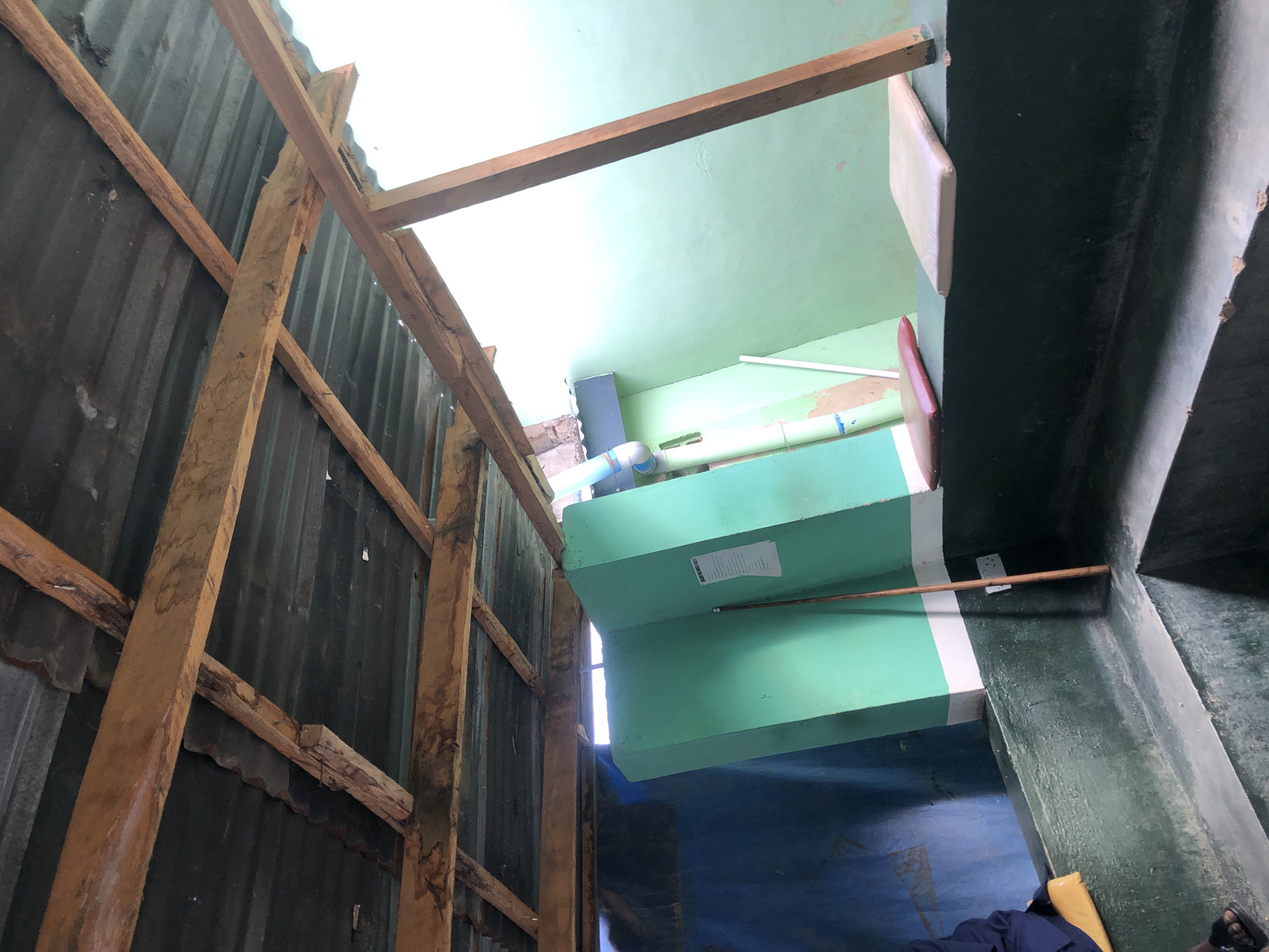 This image depicts the interior of a basic, semi-enclosed structure in Unknown, Unknown. The ceiling comprises corrugated metal sheeting supported by unpainted wooden beams, some of which exhibit dark staining. The right side of the structure is open to the exterior, admitting bright, diffuse natural light that illuminates a light green wall.
To the left, a wall is painted dark blue. A central, vertical partition, painted bright green with a white horizontal stripe, separates the blue wall from the right side. A white sheet of paper with text is affixed to this green partition. A slender wooden stick or pole leans against the partition, adjacent to a white electrical outlet with two circular sockets.
Below the partition, the structure features a multi-level concrete or masonry base. The upper ledge of this base is painted dark green, matching the lower portion of the central partition. On this dark green ledge, to the right, are two rectangular padded cushions: one beige and one reddish-brown. Below the dark green ledge, the lower levels of the base are unpainted grey concrete.
In the lower left corner, a person is partially visible, with their left foot, clad in a black sandal, resting on the concrete. A dark blue fabric (clothing) and a yellow object are also visible in this area, partially obscuring the person. A light blue pipe system runs vertically on the upper right portion of the light green wall. The overall construction appears rudimentary, utilizing readily available materials.