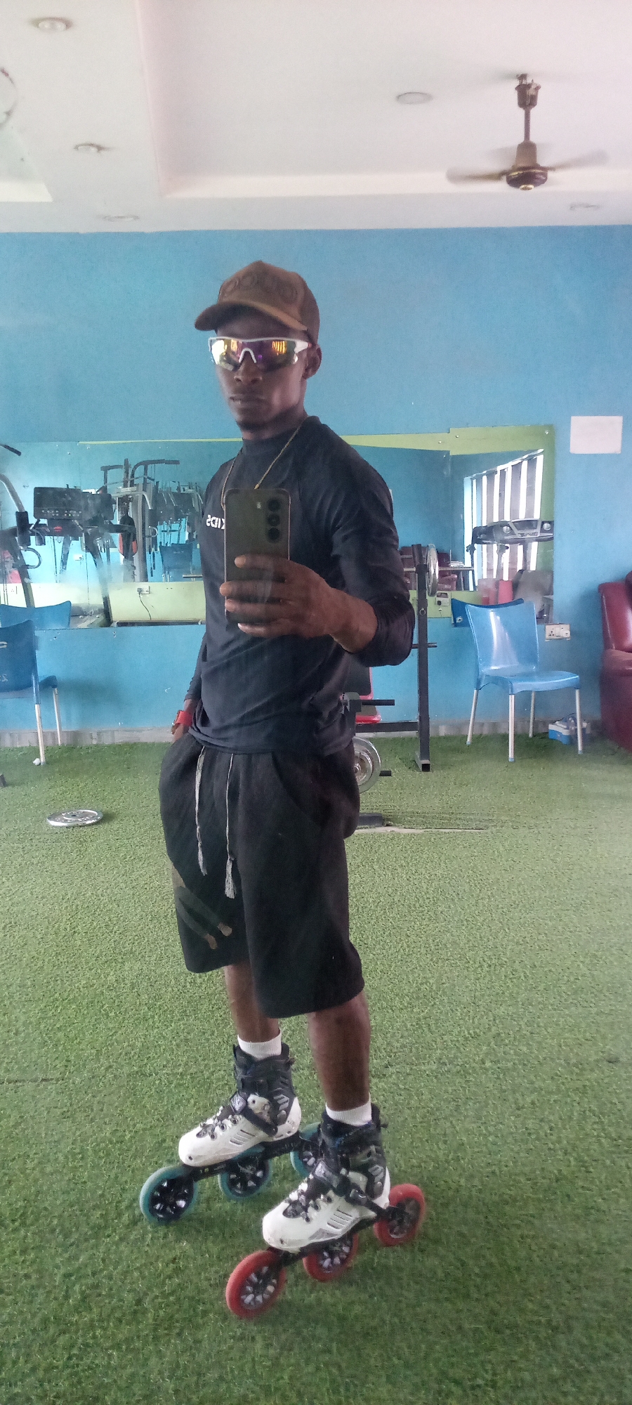 A full-body mirror selfie captures a man standing indoors, likely in a gym or training facility in Akwanga, Nigeria. He is positioned towards the left side of the frame, holding a dark-colored smartphone to take the photo. He wears a dark long-sleeved athletic shirt, black shorts with a white drawstring, white ankle socks, and prominent white and black inline skates featuring red and blue wheels. His headwear includes a brown baseball cap and reflective, mirrored sunglasses that obscure his eyes. A thin gold chain is visible around his neck.
The setting features light blue walls and a bright white ceiling with recessed lighting and a visible ceiling fan. The floor is covered with artificial green turf. In the background, reflections in the mirror show various gym equipment, including weight machines and free weights. Blue plastic chairs and a red upholstered seat are also visible in the reflections. The mirrored surface on the back wall reflects reversed text, possibly "KIDS" or "2013". The man's pose is confident and casual, suggesting he is either preparing for or concluding a skating session.