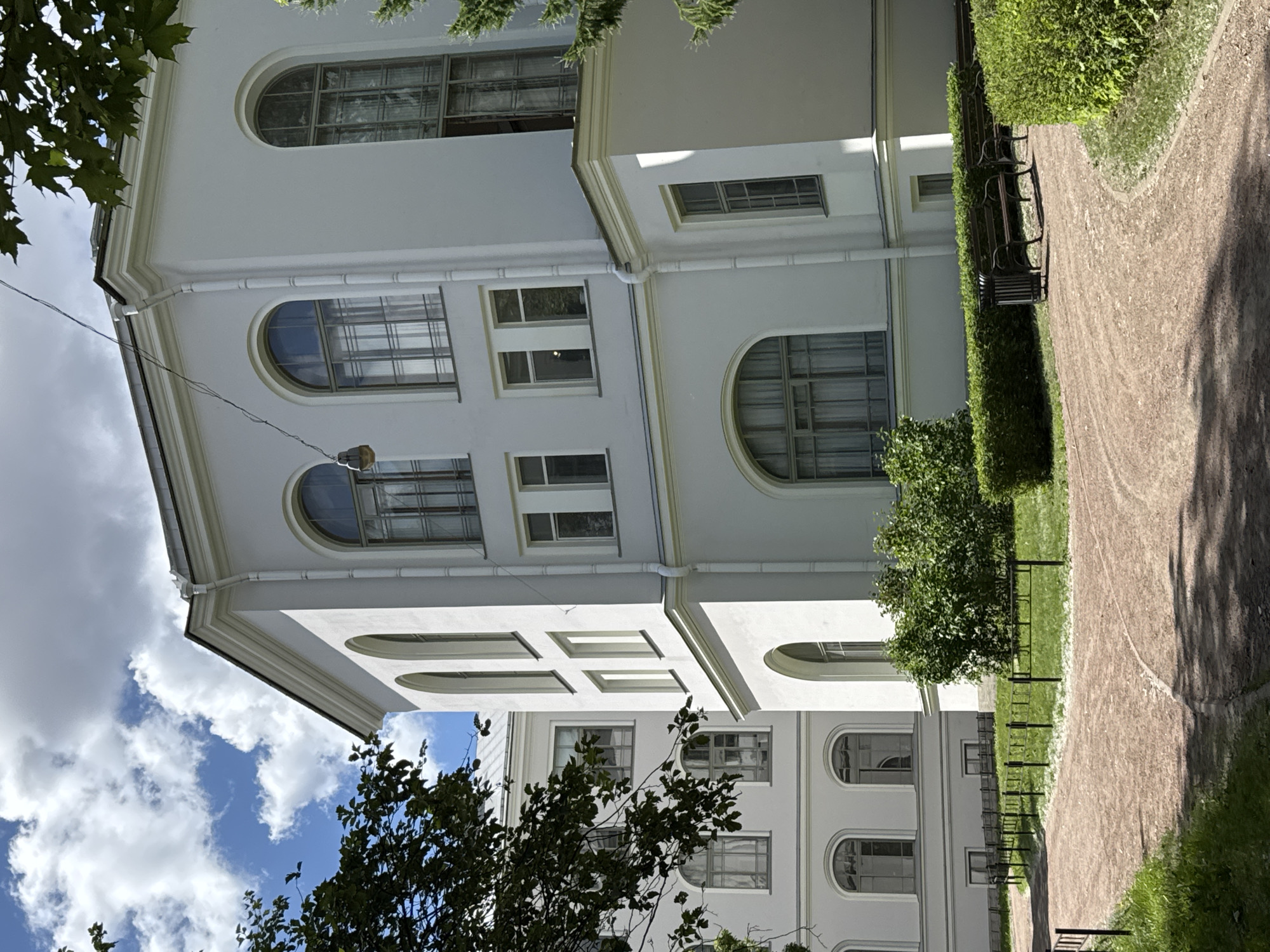 This media file depicts the exterior of a large, multi-story white building with a neoclassical architectural style, likely a public institution or historic building. The building features numerous windows, many of which are arched and divided into multiple panes. Some windows appear to have reflective glass. The facade is accented with decorative molding.
In the foreground and to the right, a gravel path curves through a manicured park-like setting. The path is bordered by neatly trimmed green hedges and grass. Several dark metal benches with wooden slats are visible along the path, suggesting a place for public recreation or rest. Lush green trees and foliage frame the building and the park, with some leaves casting shadows on the path.
The scene is outdoors, under a bright sky with scattered white clouds, indicating daytime. The lighting suggests it is a clear and pleasant day. The orientation of the image is rotated, with the sky appearing on the left and the building and path on the right.
There is no visible text in the image. The overall impression is one of calm and ordered public space, characteristic of a well-maintained historical urban environment. The presence of benches and the park setting suggest a location where people might gather or stroll. The architectural style and the greenery hint at a location in Saint Petersburg, Russia, known for its grand buildings and parks.