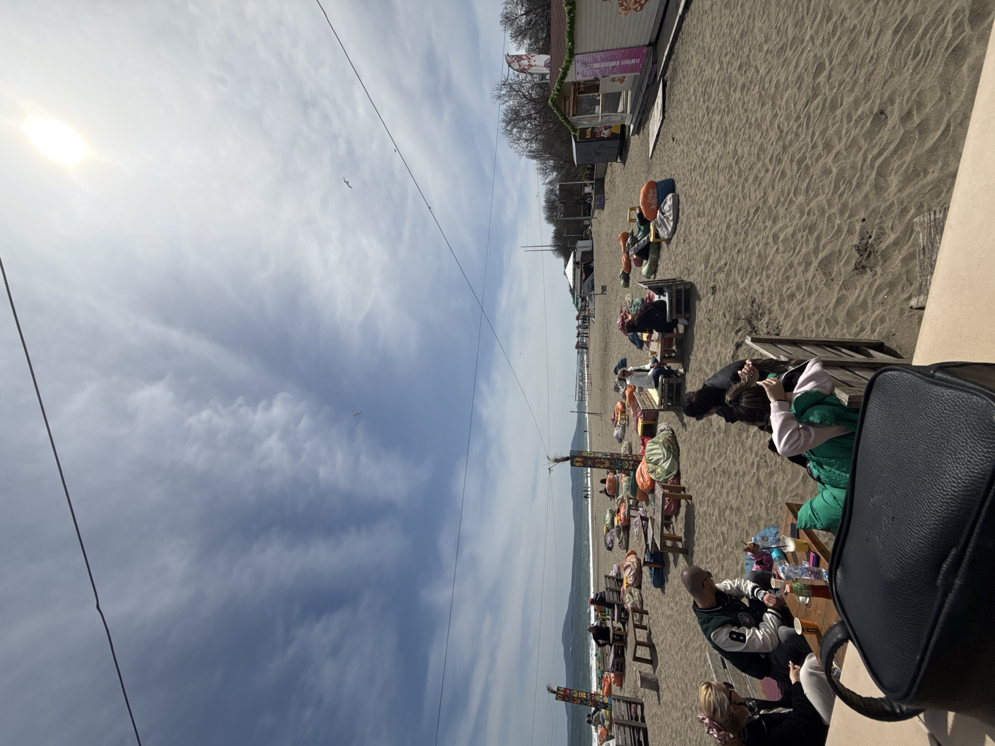 This image captures a vibrant daytime scene on a sandy beach in Burgas, Bulgaria, featuring people relaxing at a casual beachside establishment. The perspective is slightly elevated, looking across the beach towards the sea and horizon.
In the immediate foreground, on the right side, a black shoulder bag rests on a light-colored surface. Nearby, people are seated at a wooden table. One individual with blonde hair is partially visible, while another wears a white and green letterman-style jacket with the number '8' on the sleeve. Plastic cups and bottles, along with other small items, are on their table.
Further into the midground, numerous individuals are scattered across the beach, sitting at wooden tables and chairs on the sand. Many of these chairs and tables are adorned with colorful cushions or fabric coverings. Bright orange inflatable objects, one clearly marked 'VERO SPORTS', are placed near some seating arrangements. Decorative poles, painted with colorful patterns, are also spaced along the beach. The sand shows various patterns and footprints.
Towards the left side of the scene, the gentle waves of the sea meet the shore, and a distant coastline can be seen faintly in the hazy background. Along the back of the beach, behind the seated people, a series of simple wooden stalls or kiosks are visible. Some of these structures are decorated with garlands and banners, displaying text that is largely illegible, though some pink and white lettering can be discerned on one. Behind the stalls, the bare branches of trees suggest either early spring or late autumn.
The sky occupies the upper portion of the image, showing a bright, almost glaring sun in the upper left corner. The sky is partly cloudy with patches of blue, and scattered white clouds stretch across. A thin black wire runs diagonally across the upper part of the sky. Several small birds are in flight against the cloudy backdrop.
The overall atmosphere is one of relaxation and leisure, with people enjoying the pleasant weather by the sea.