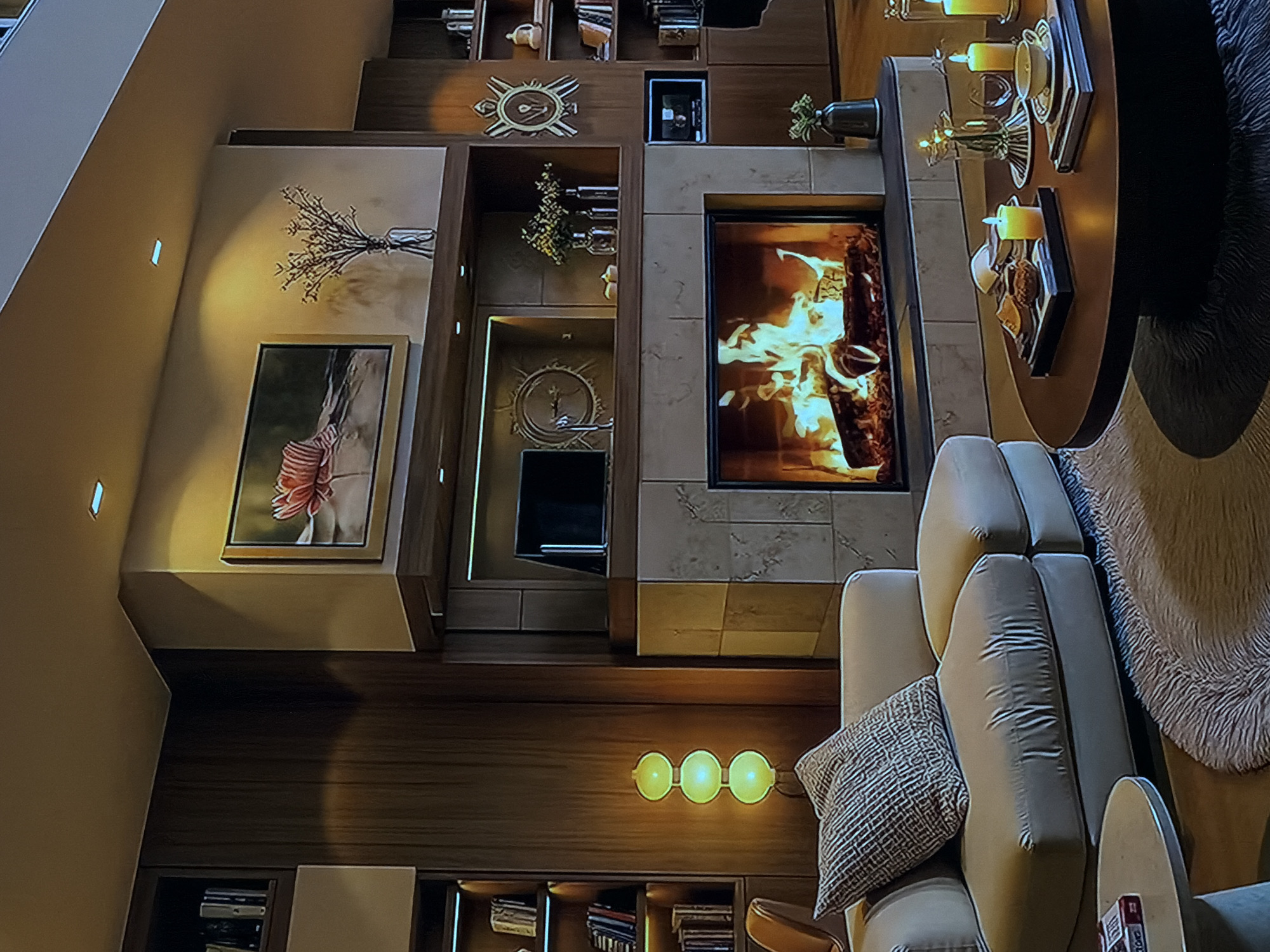The image displays a modern and luxurious living room interior, likely within a residential setting in Sofiivska Borshchahivka, Ukraine. The scene is captured from a tilted, elevated perspective, showcasing a multi-level or open-plan design with prominent architectural details.
The focal point is a contemporary fireplace featuring a wide glass front, behind which lively flames dance around logs, casting a warm, orange glow. The fireplace is framed by light-colored stone tiling and integrated into an extensive system of dark wooden built-in shelving and paneling that covers the walls. These shelves are adorned with various decorative objects, including two stylized clocks with metallic accents, vases holding dried branches or foliage, and other small ornaments.
To the left of the fireplace, a section of the wall features a framed artwork depicting a large, abstract pink or red flower. Below this, further dark wood paneling extends, incorporating additional shelving units that appear to hold books or display items, some of which are illuminated. A wall-mounted sconce with three circular, glowing yellow lights provides additional ambient lighting in this area.
The seating arrangement consists of plush, light-colored sofas or armchairs, adorned with soft cushions and a patterned throw pillow, inviting relaxation. Adjacent to the seating, a round, dark-colored coffee table holds multiple lit candles placed on decorative trays, alongside other small items, contributing to a cozy and intimate atmosphere. A fluffy, light-colored rug is partially visible beneath the table and seating.
The room is illuminated by a combination of recessed ceiling and wall lights, the glow from the fireplace, and the sconce, suggesting an evening or nighttime setting. The overall aesthetic is one of warmth, sophistication, and comfort, with a strong emphasis on natural materials like wood and stone. No people are visible in the image, and no discernible text can be read on any objects.