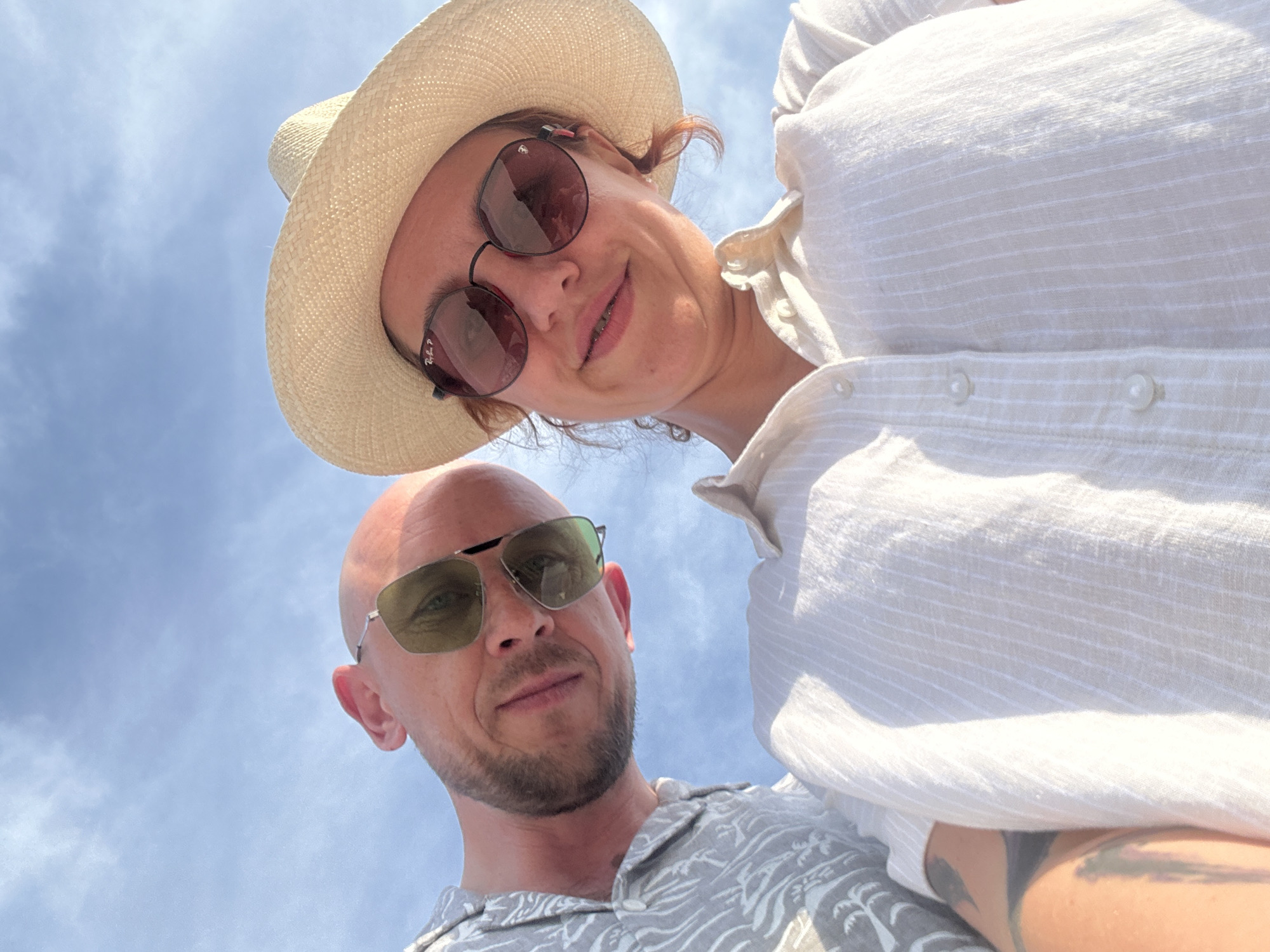 An extreme low-angle outdoor photograph depicts two individuals, one male and one female, looking directly downwards into the camera lens. The background consists solely of a clear, light blue sky with very faint, sparse white cloud formations, indicating bright daylight conditions. This scene is identified as taking place in the city of Sochi, Russia.
The male subject, positioned on the left side of the frame, has a bald head, light skin, and a short, reddish-brown beard. He wears sunglasses with reflective green-gold lenses in a thin silver frame. He is dressed in a light grey short-sleeved collared shirt with a subtle pattern of lighter grey tropical foliage or palm trees. His head is tilted slightly, and he displays a mild smile.
The female subject, on the right, wears a wide-brimmed straw hat of a natural, light beige color. She wears round, dark-lensed sunglasses; the brand "Ray-Ban P" is visible on the left lens. Her reddish-brown hair is visible beneath the hat. She is attired in an off-white or light beige short-sleeved collared button-up shirt with subtle vertical stripes. She is smiling broadly, revealing dental braces on her upper teeth. A dark, abstract tattoo is partially visible on her left upper arm or shoulder.
Both subjects are leaning into the frame from above, appearing in close proximity to each other. Their direct gaze at the camera suggests they are posing for a photograph.